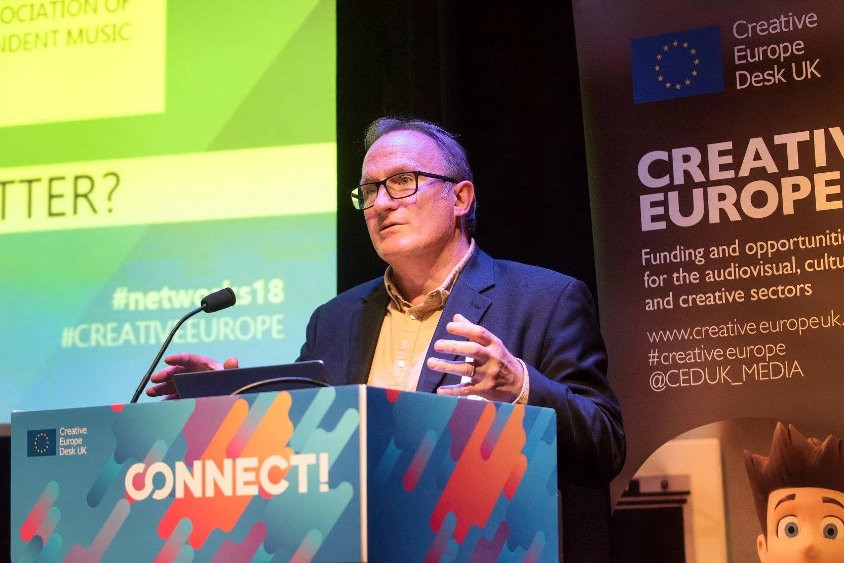 David Drake with glasses and gray hair speaking at a podium during a European conference. The background shows a large screen and a banner with the European Union flag and the words 'Creative Europe' and related information.