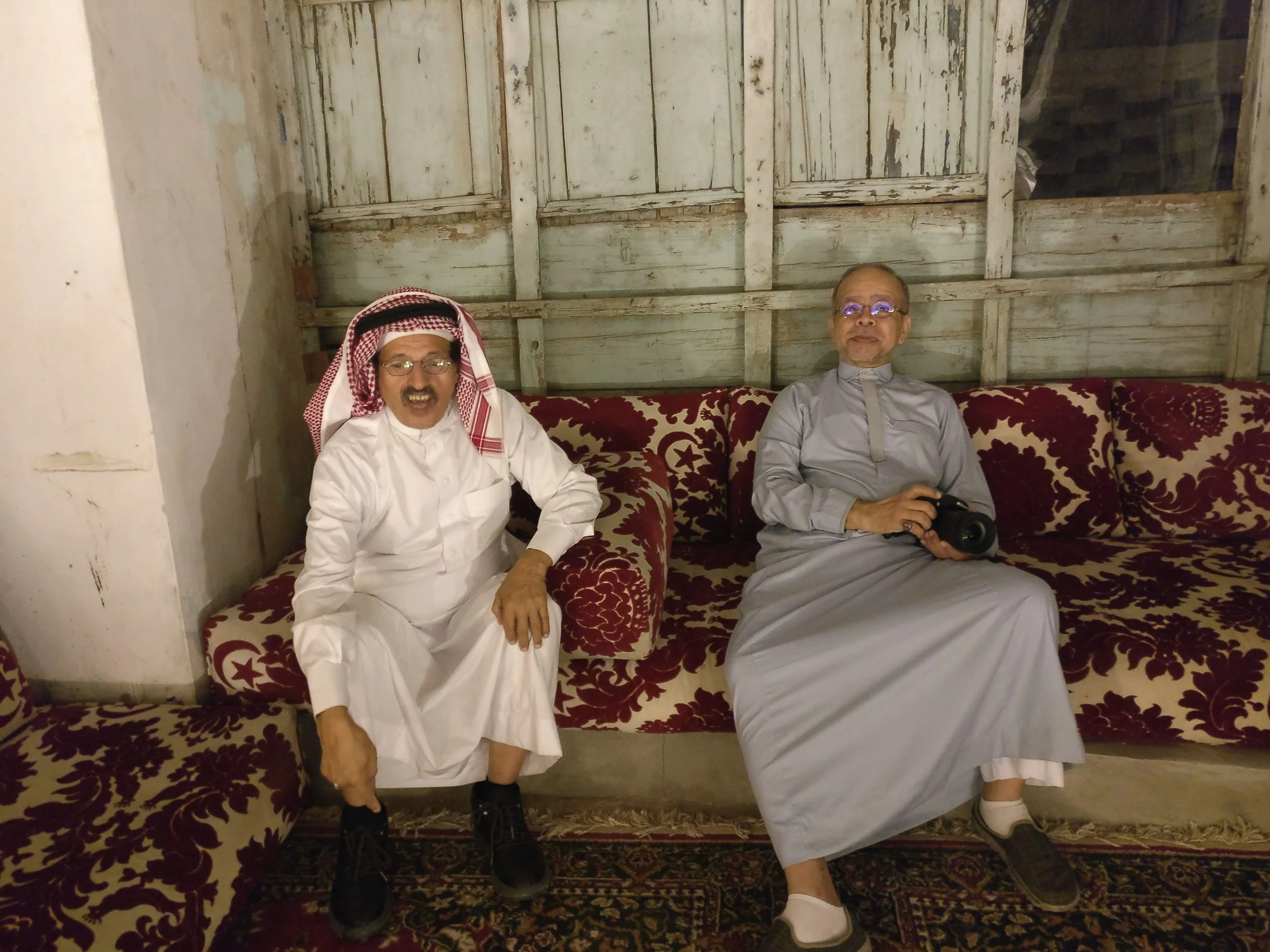 Two men sitting on a patterned sofa in a rustic room. The man on the left is wearing traditional Middle Eastern attire, including a white thobe and a red and white checkered keffiyeh. The man on the right is dressed in gray traditional clothing and i