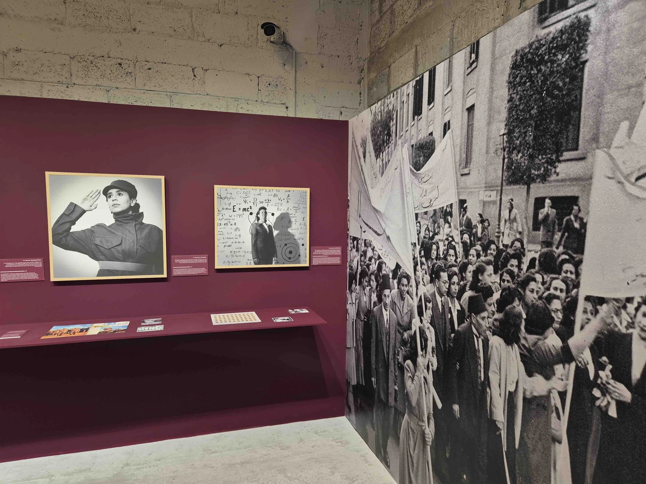 Exhibition display featuring black and white photographs of women, including a woman saluting in military uniform and a woman standing in front of surfaces with mathematical formulas, with a large crowd photograph on a wall to the right.