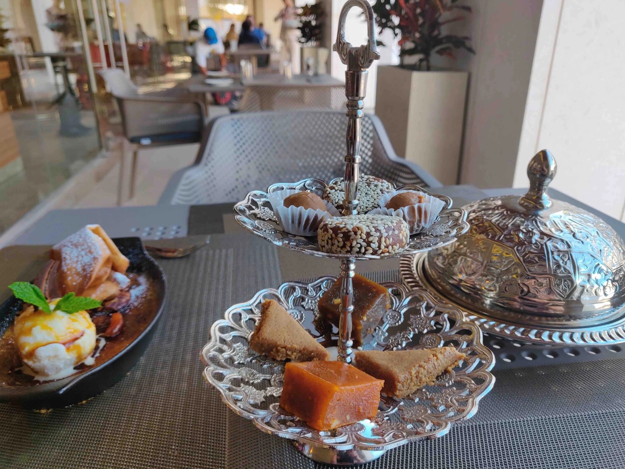 A table set with a silver platter of assorted desserts, including cookies with sprinkles, pieces of cake, and other confections, with a black plate of desserts garnished with a mint leaf and powdered sugar in the foreground, in a well-lit restaurant 