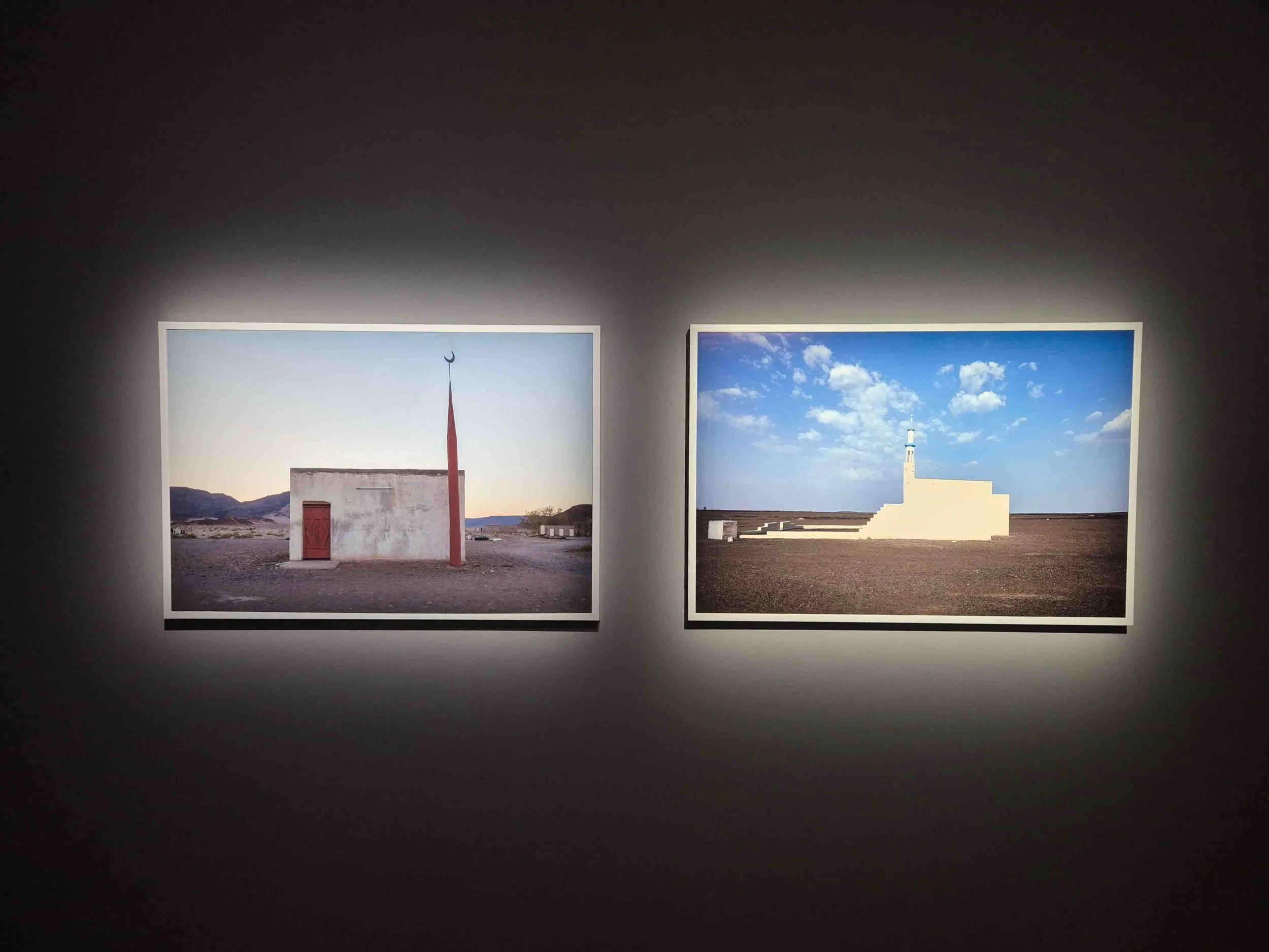 Two framed photographs by Moath Alofi of minimalist structures on a dark gallery wall, one featuring a small building with a red door and a tall red pole, the other showing a white monument with a lighthouse on top under a partly cloudy sky.