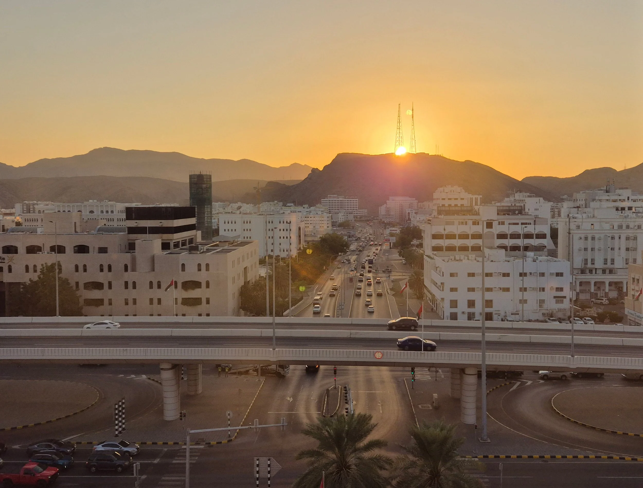 Sunset over Muscat, Oman,  with white buildings, a highway with cars, and mountains in the background.