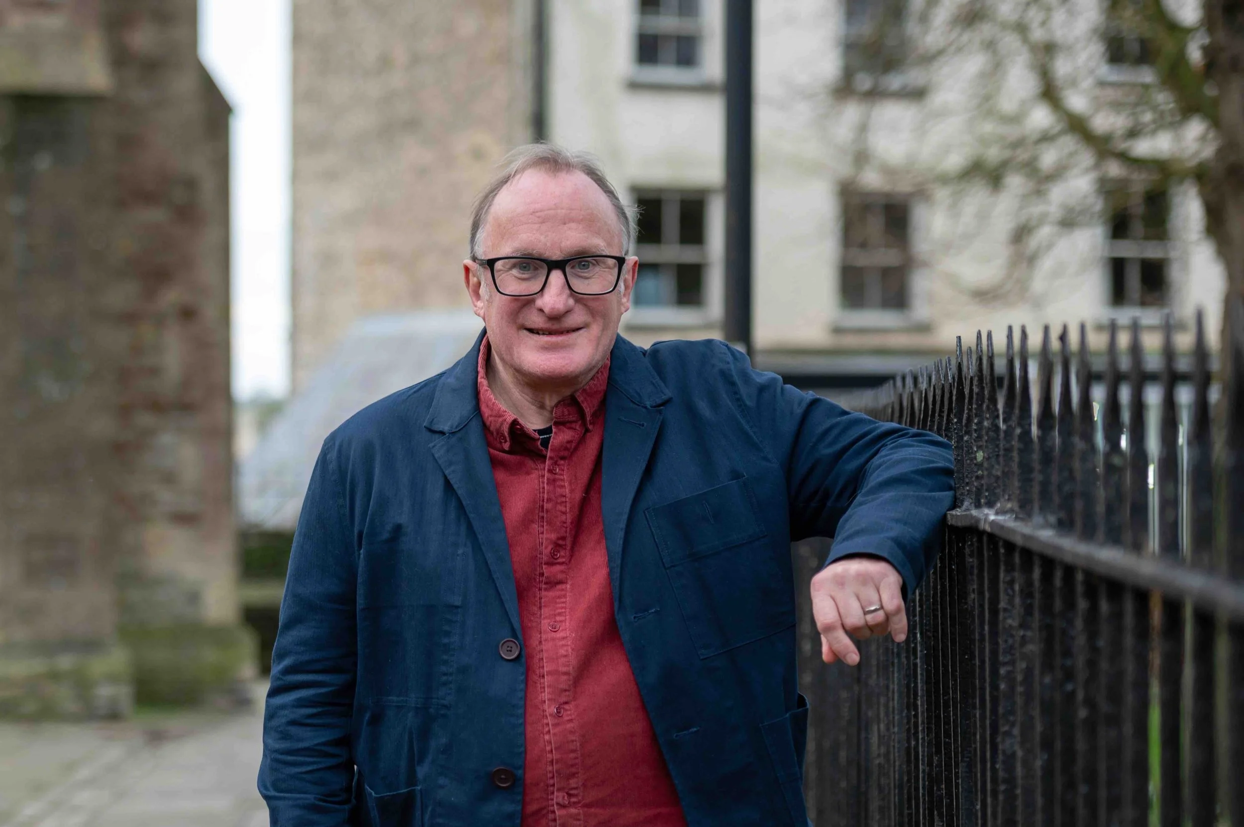 A middle-aged man with glasses and short, light-colored hair smiling outdoors. He's wearing a red shirt and a navy jacket, leaning on a black metal fence with houses and trees in the background.