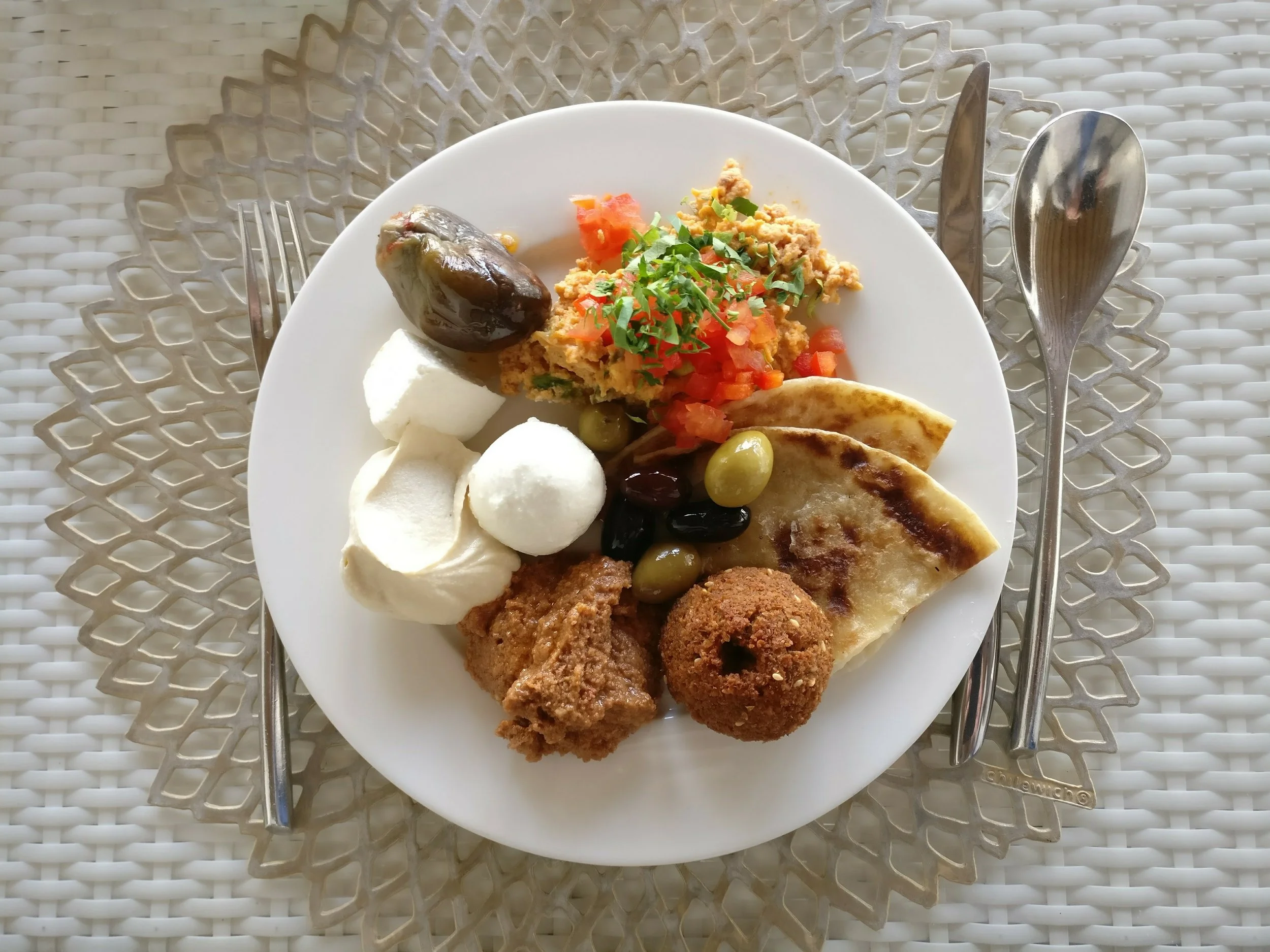 A white plate of assorted Middle Eastern appetizers and foods, including falafel, olives, hummus, pita bread, stuffed grape leaf, and tomato and parsley garnish, on a woven placemat with a fork and spoon on either side.