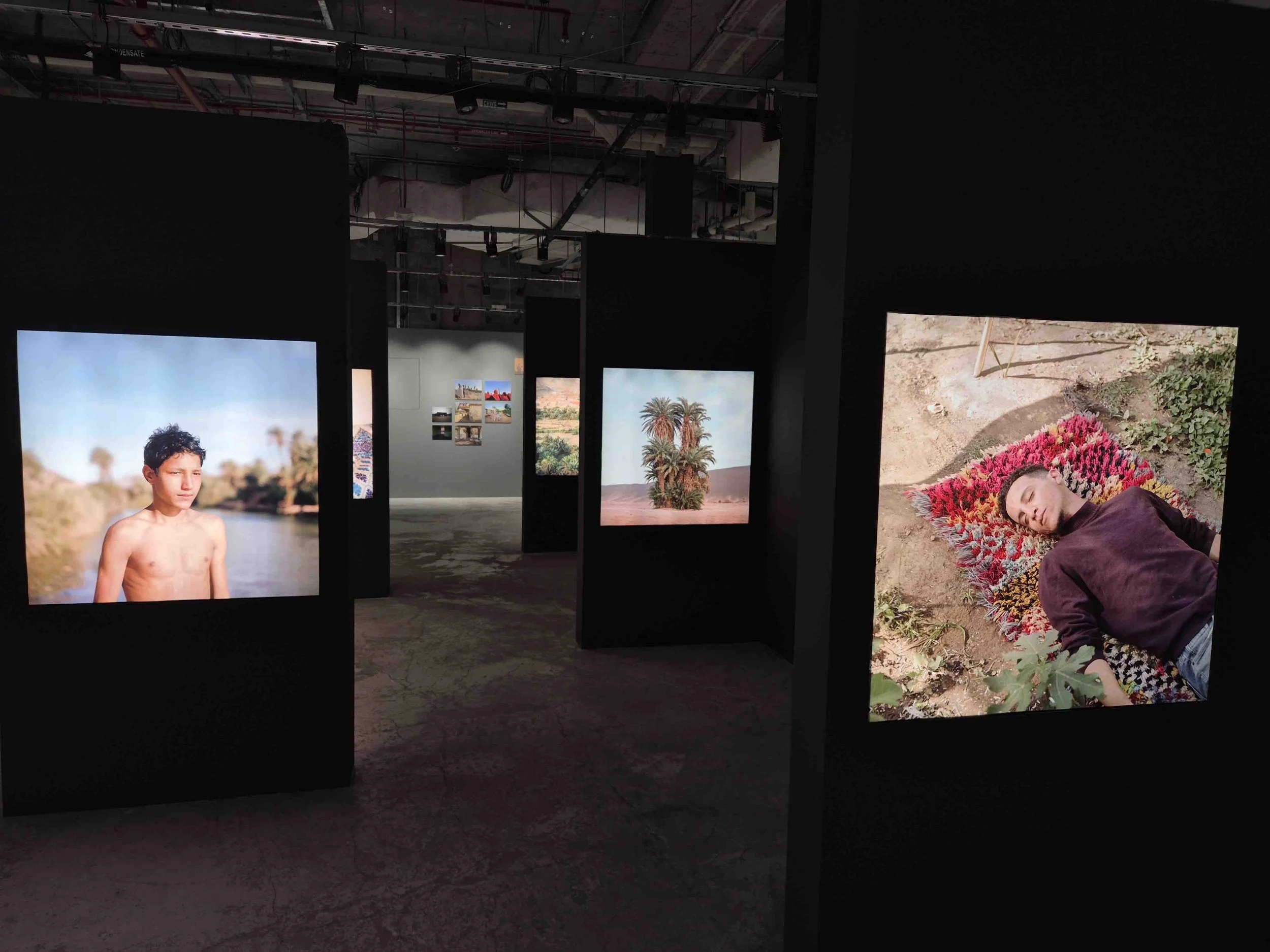 An art gallery display with several framed photographs by Mohammed Kilito, including a shirtless boy near a river, a person lying on a colorful rug, and palm trees in a desert landscape.