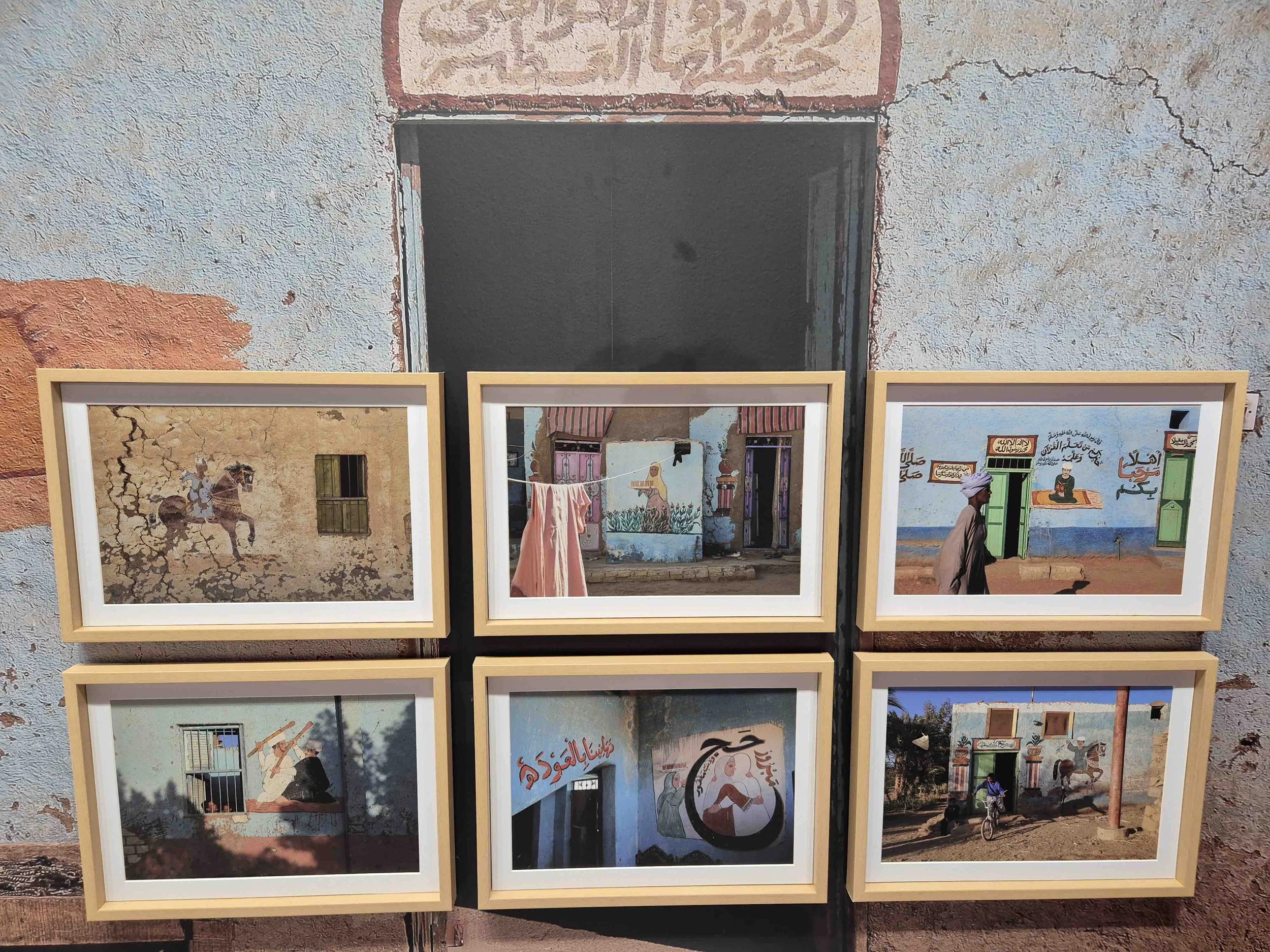 Egyptian artist Wafaa Samir's  photographs of murals on walls, depicting scenes of people, animals, and Arabic calligraphy, set against a textured, weathered wall with an open doorway.