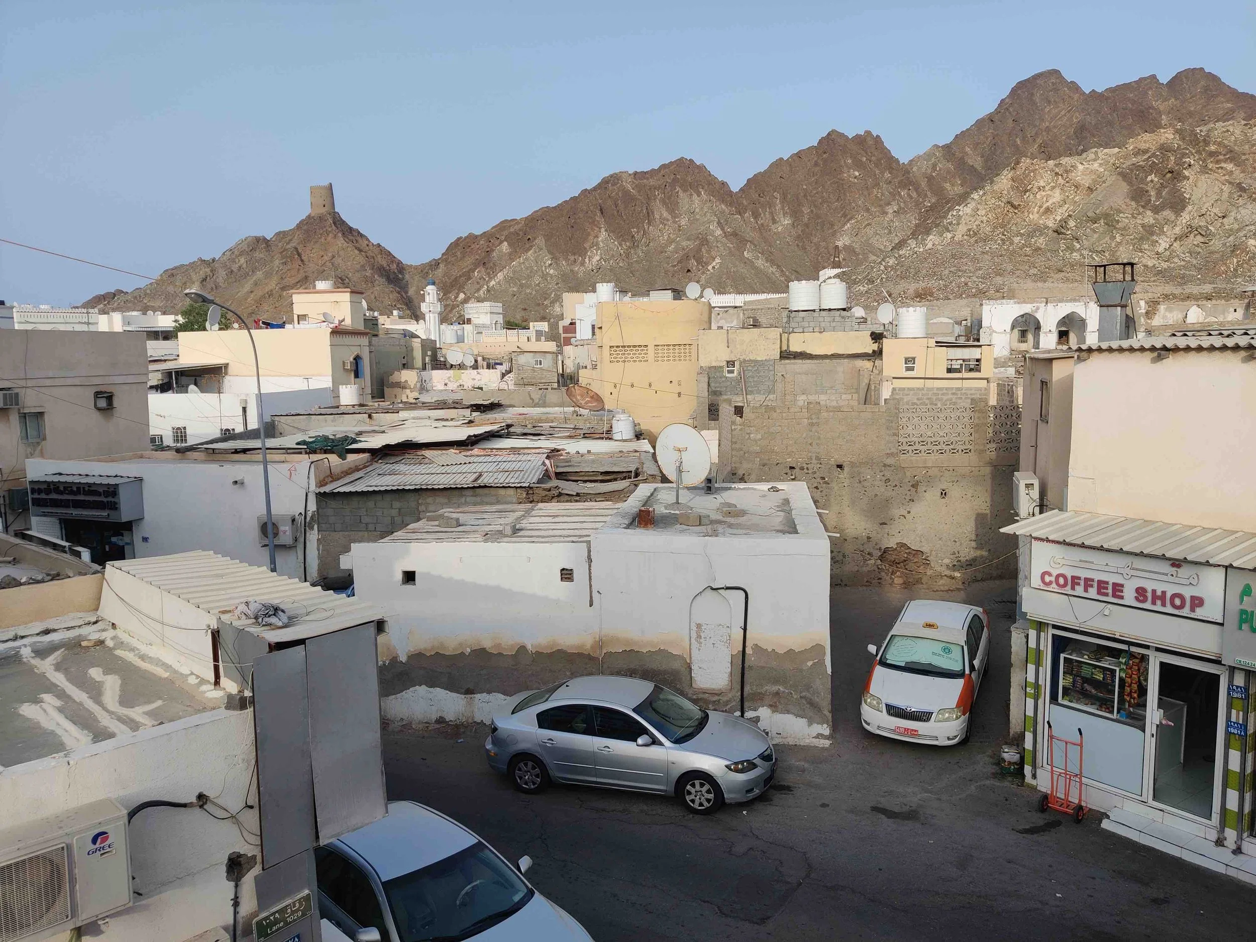 Muttrah in Oman with white and beige buildings at the foot of rocky mountains. There is a coffee shop on the right, several parked cars, and satellite dishes on rooftops.