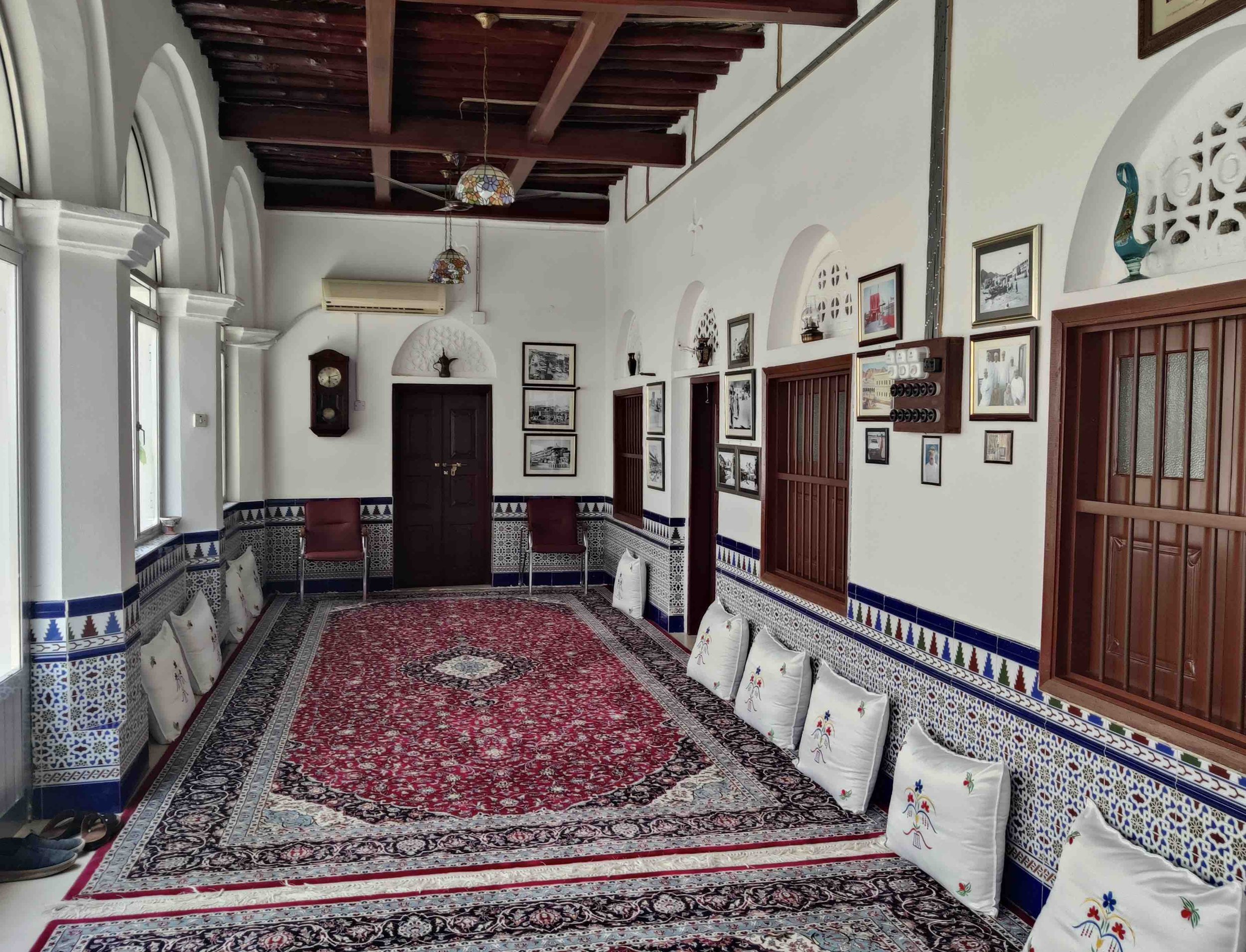 Interior of a traditional room with white walls, arched windows, and wooden accents. It features a decorative red and black patterned rug, cushioned seating along the walls, and framed artwork. The ceiling has exposed wooden beams and hanging glass l