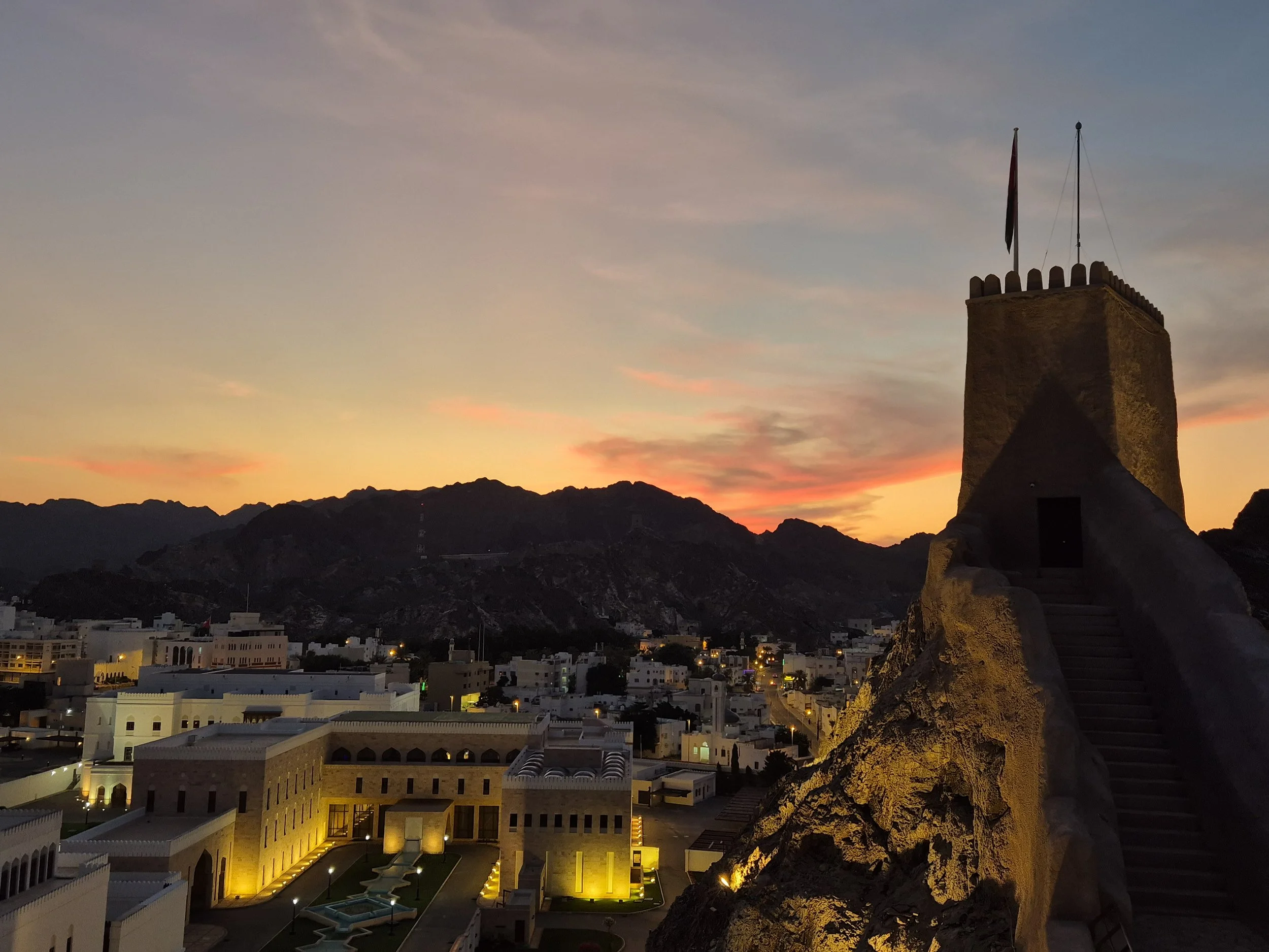 Old Muscat cityscape at sunset with a historic tower on a rocky hill in the foreground and mountains in the background, illuminated buildings, and a colourful sky.