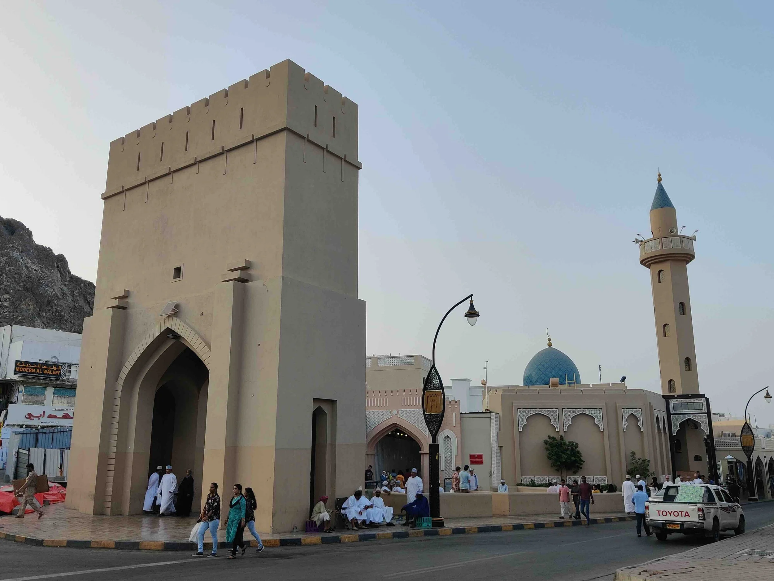 The city gate in Muttrah, Oman, with a blue dome and tall minaret, behind it, and people walking and sitting outside, on a street with cars and streetlights.