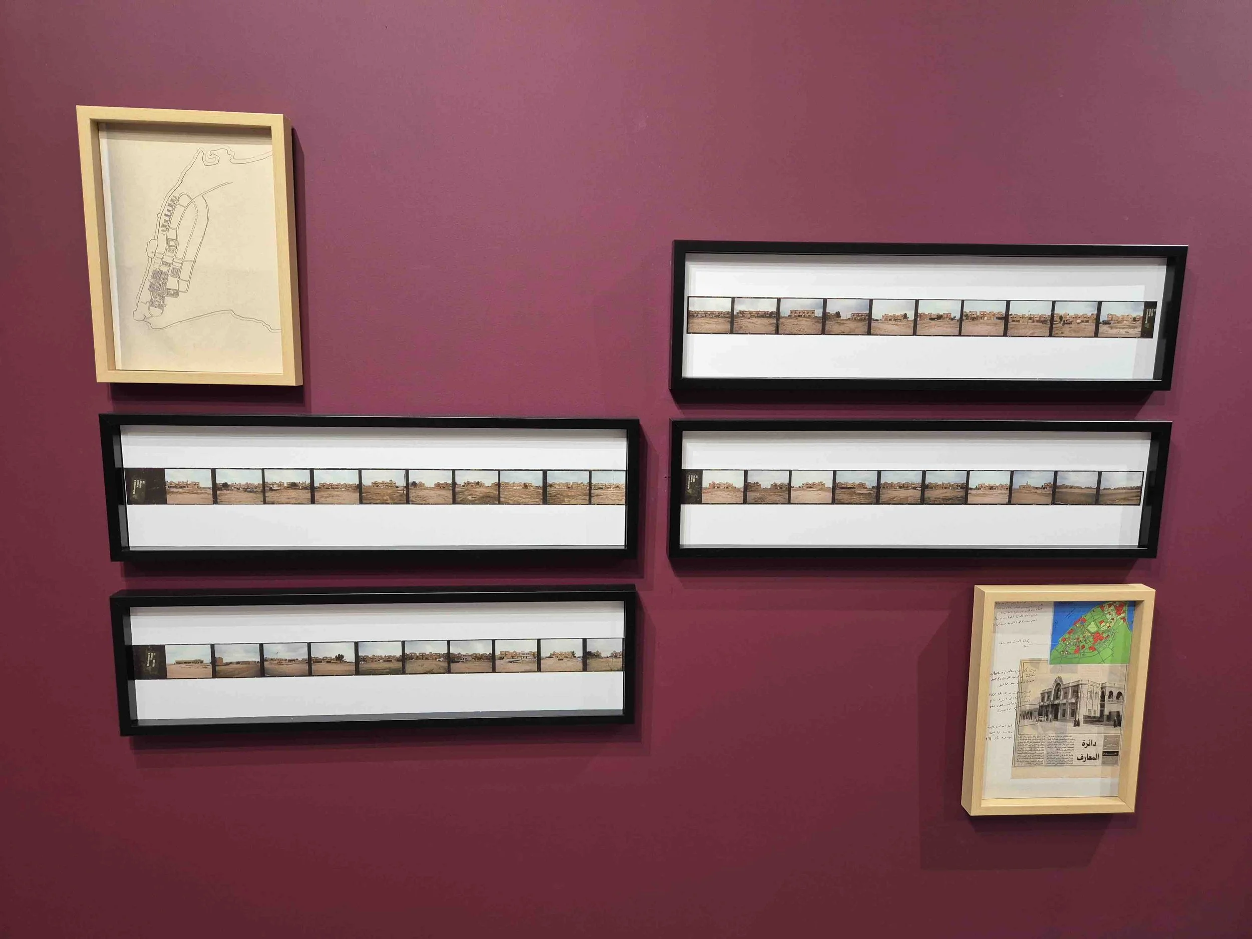 Display of panoramic photographs by Mohammed AlKour on a purple wall with two framed maps or artwork.