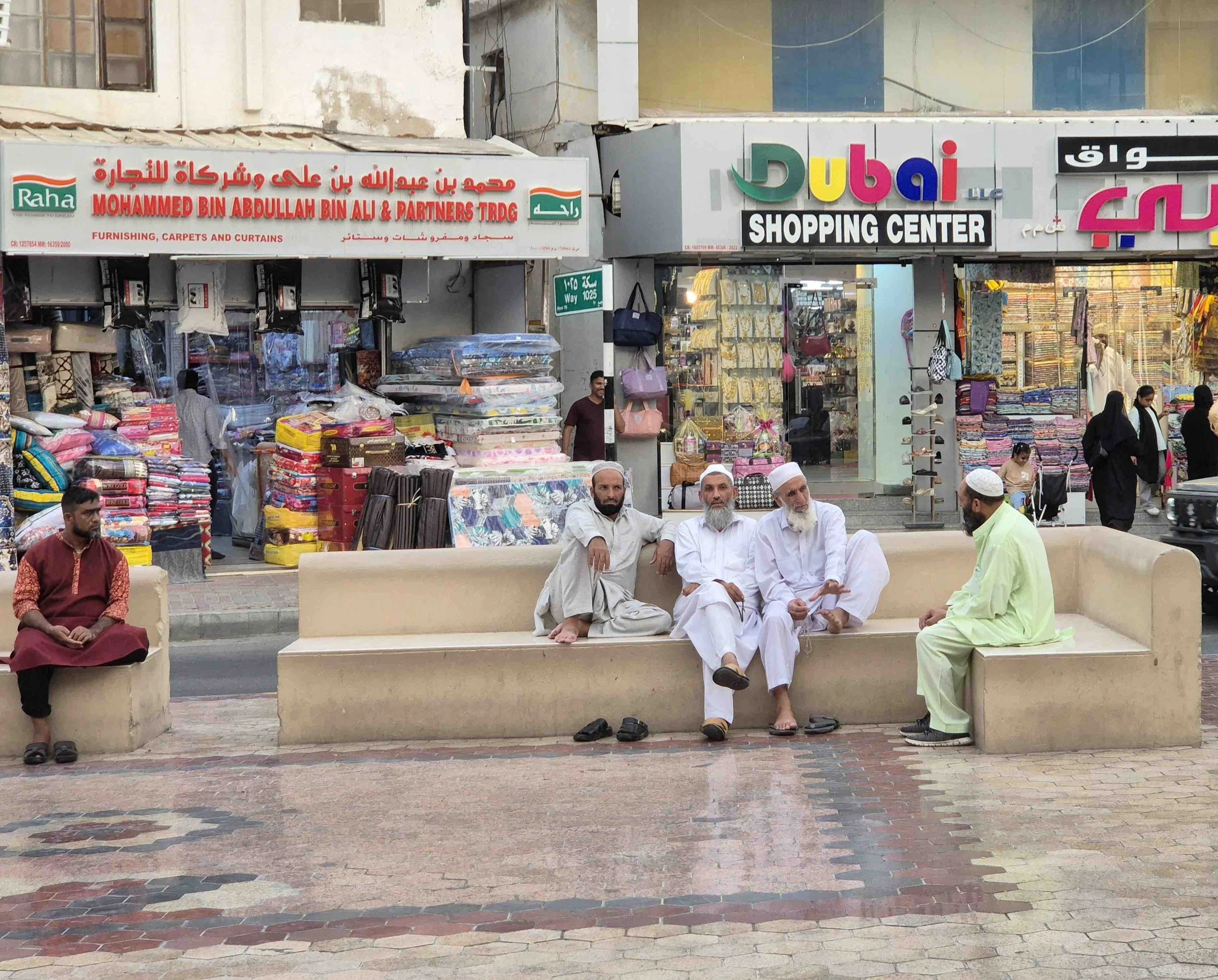 Four men sitting on a bench in a busy outdoor market in Muttrah, Oman, , with shops selling textiles and bags in the background.