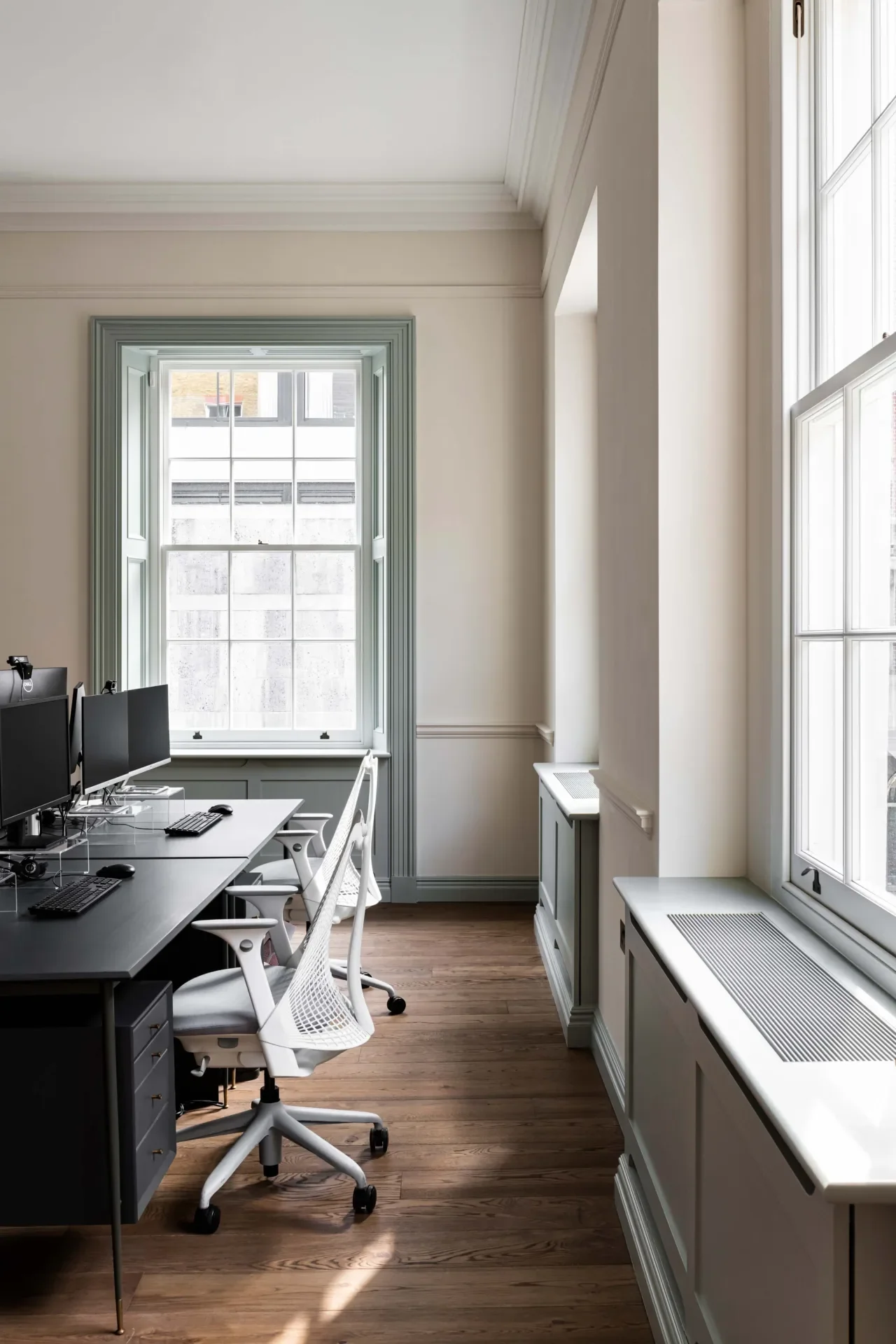 Interior of an office with white walls, large windows allowing natural light, a black desk with three computer monitors, white ergonomic chairs, and wooden flooring.