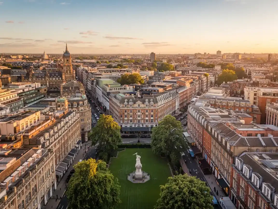 Aerial view of a city neighborhood with historic buildings and a green park with a statue at the center, during sunset.