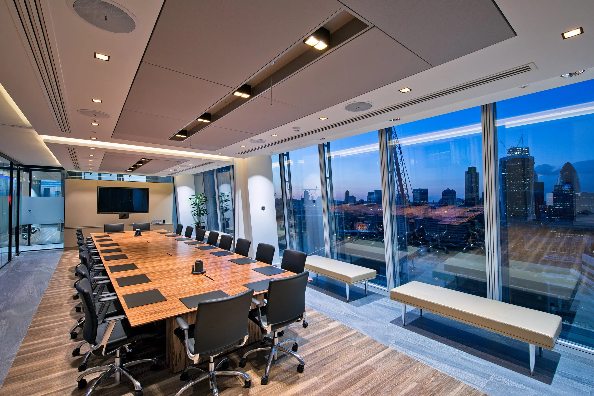 Modern conference room with large window view of cityscape at dusk, a long wooden table with black chairs, a large flat-screen TV on the wall, and a few potted plants.