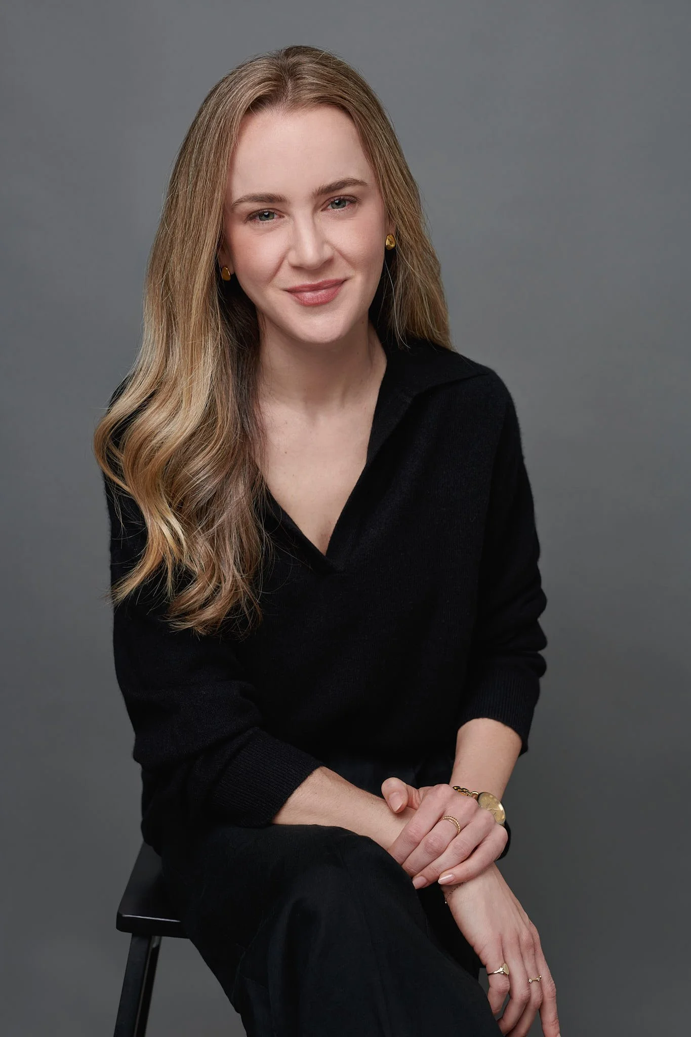 A woman with long, wavy blonde hair wearing a black long-sleeved top and gold jewelry sitting on a black chair against a plain gray background.