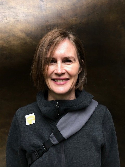A woman with short light brown hair smiling, wearing a black jacket with a gray strap across her chest, standing in front of a dark, metallic background.
