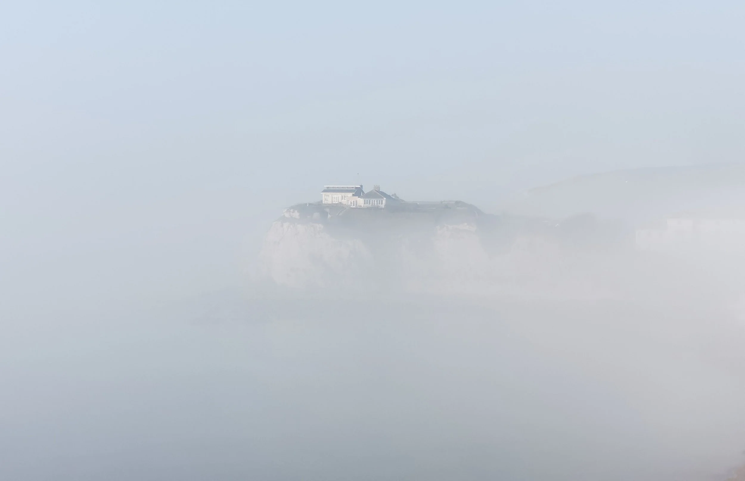 A house situated on a cliff surrounded by thick fog, with the ocean and sky in the background.