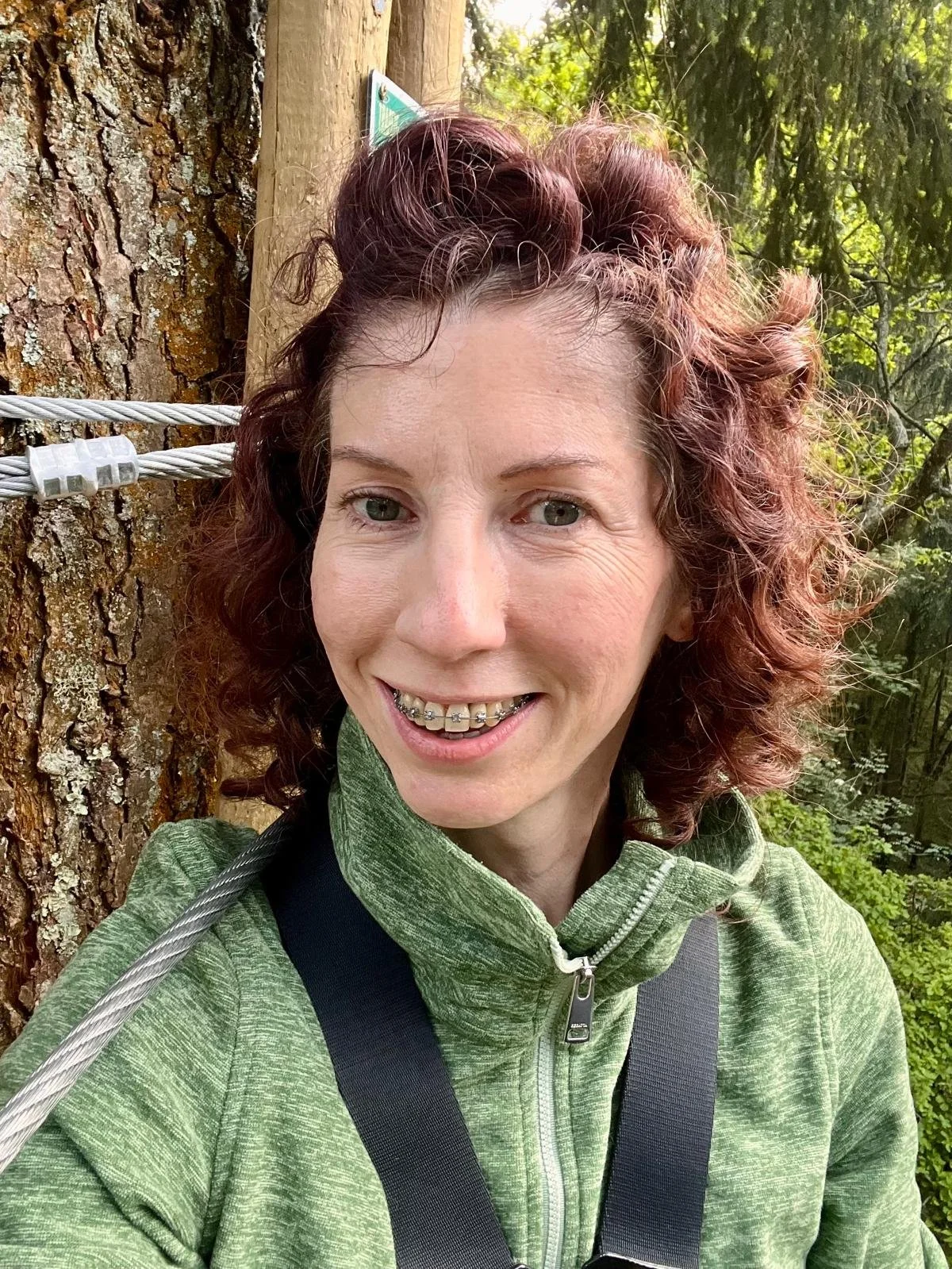 A smiling woman with braces and curly reddish-brown hair taking a selfie outdoors in a wooded area, standing near a tree with a safety cable.