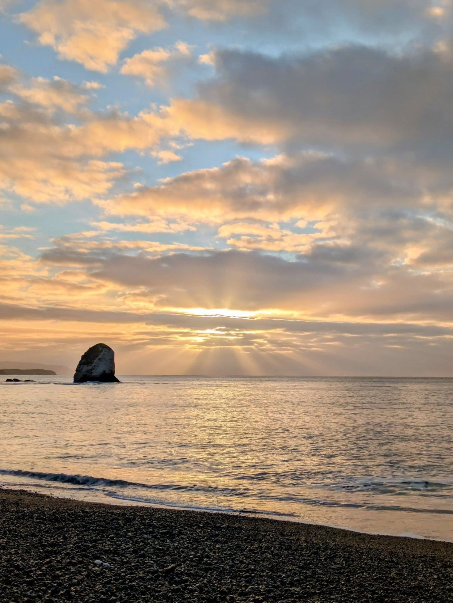 Sunset over the ocean with a large rock formation in the water and a pebble beach in the foreground.