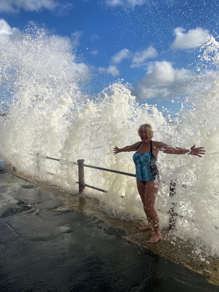 Older woman standing on a concrete surface near the ocean with big waves crashing behind her, smiling and holding out her arms.
