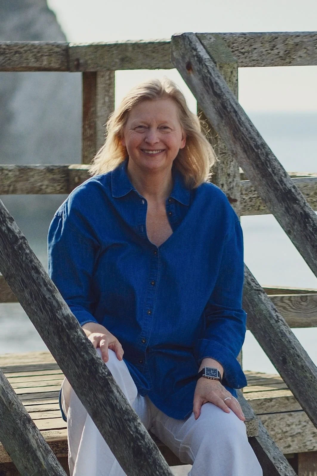 A woman with blonde hair smiling, wearing a blue shirt and white pants, sitting on a wooden dock.
