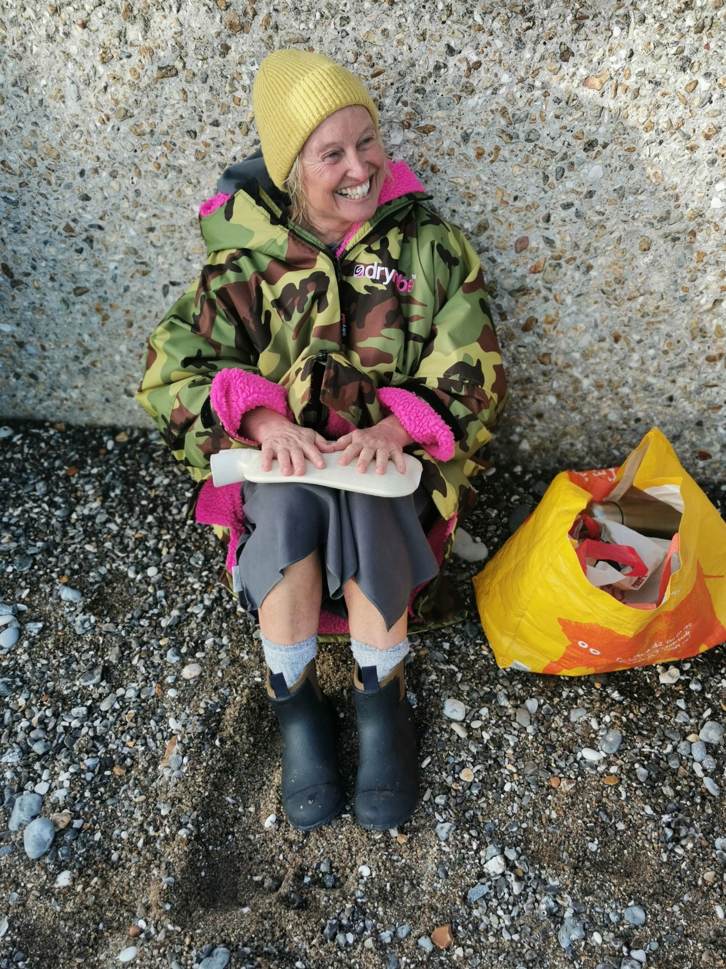 A smiling woman in a yellow beanie, camouflage jacket with pink accents, and black rubber boots sitting on a pebble beach, with a yellow shopping bag nearby.