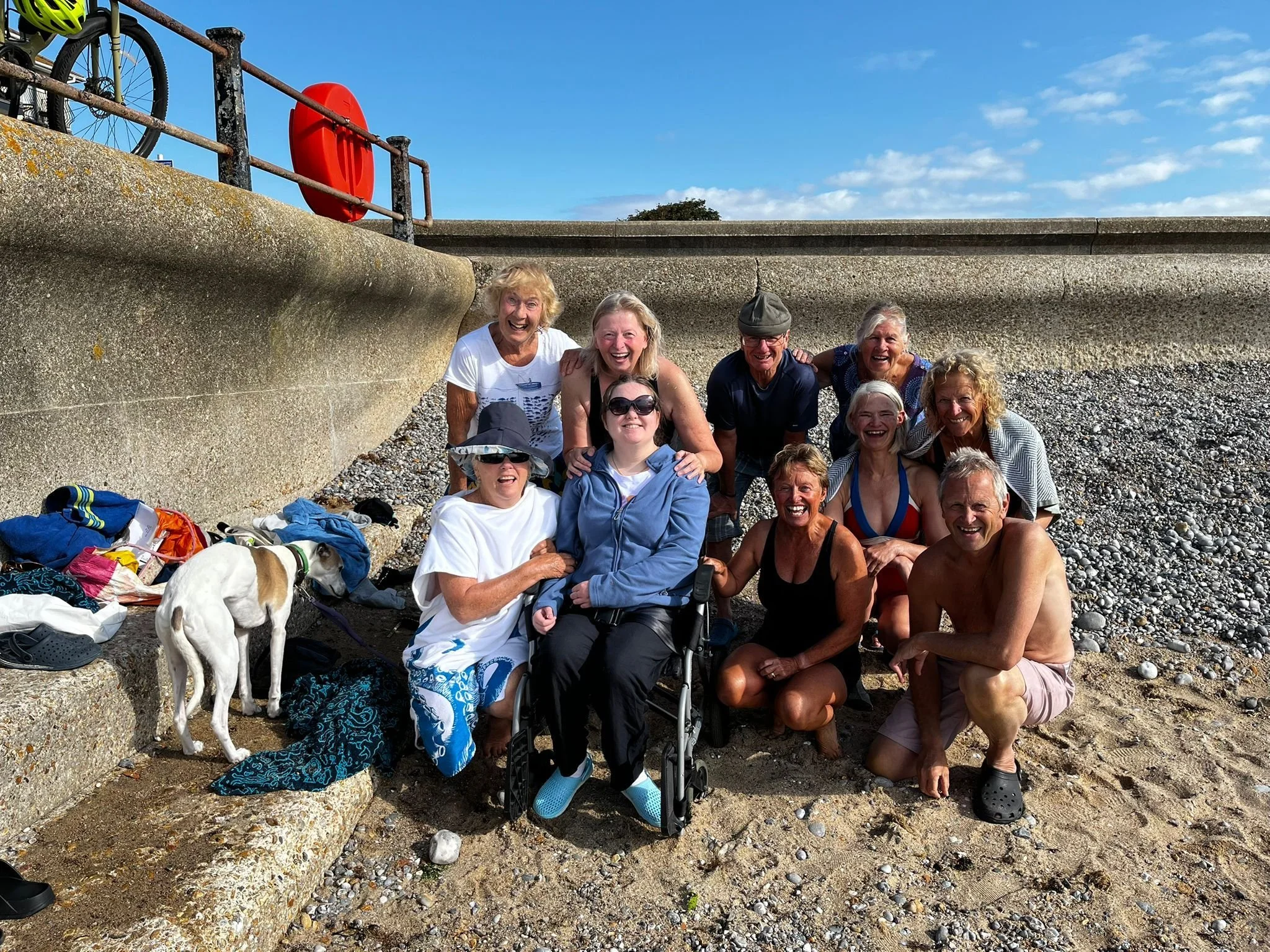 Group of adults and children sitting on a pebble beach, smiling for the camera, with clothes and backpacks scattered around. A dog stands near the front, and the background features a low concrete wall, a railing, a red lifebuoy, a bicycle, and a blu