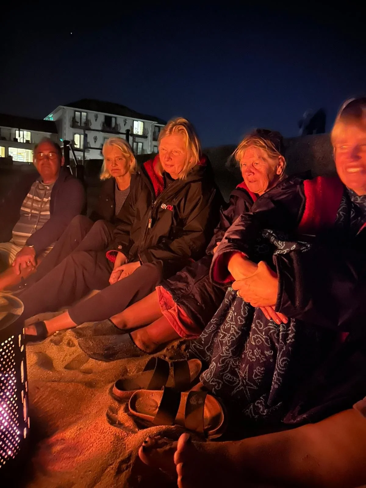 People sitting around a campfire outdoors at night, with a building and dark sky in the background.