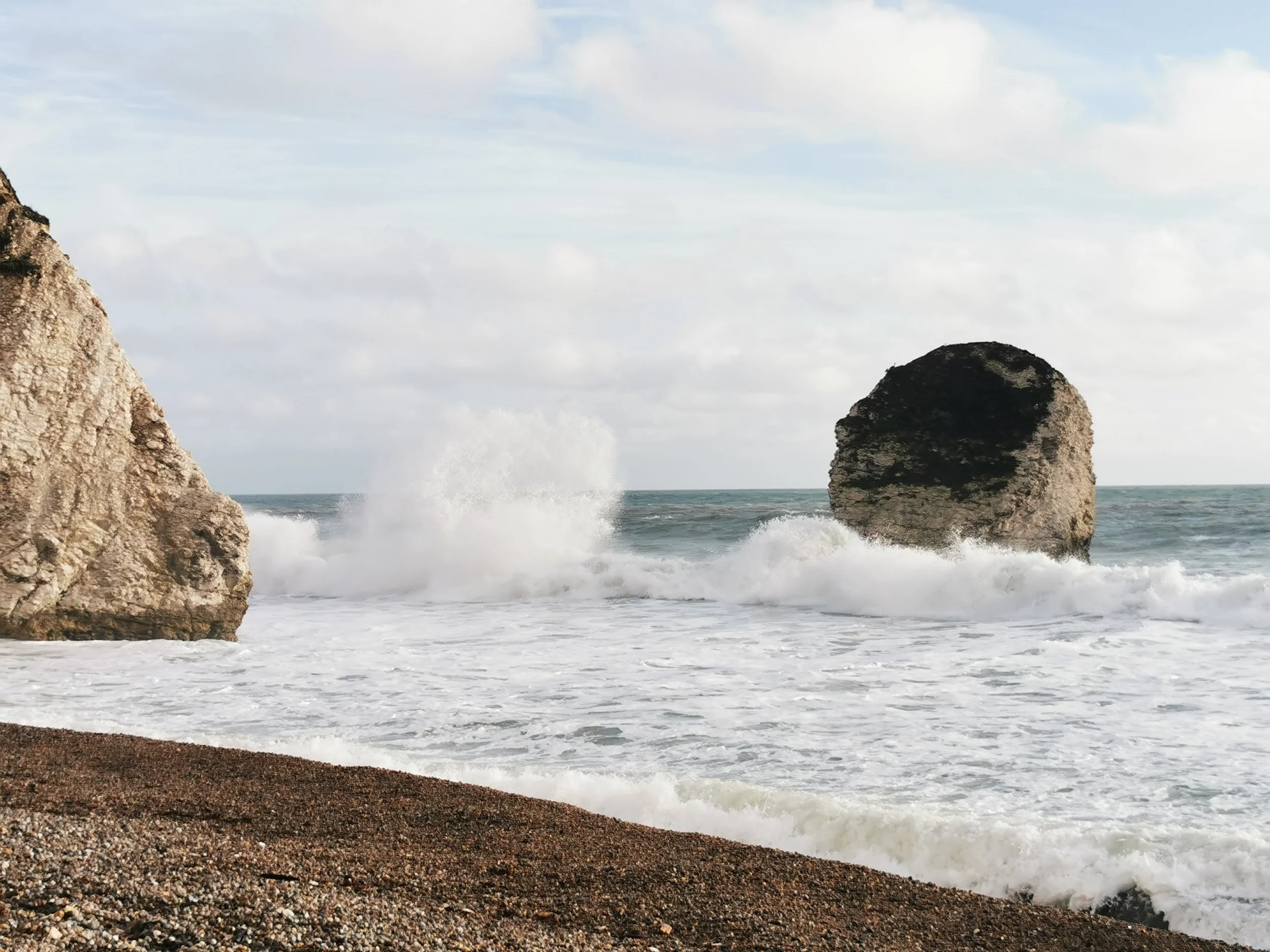 Cliffs and large rocks by the ocean with waves crashing against them and cloudy sky above.