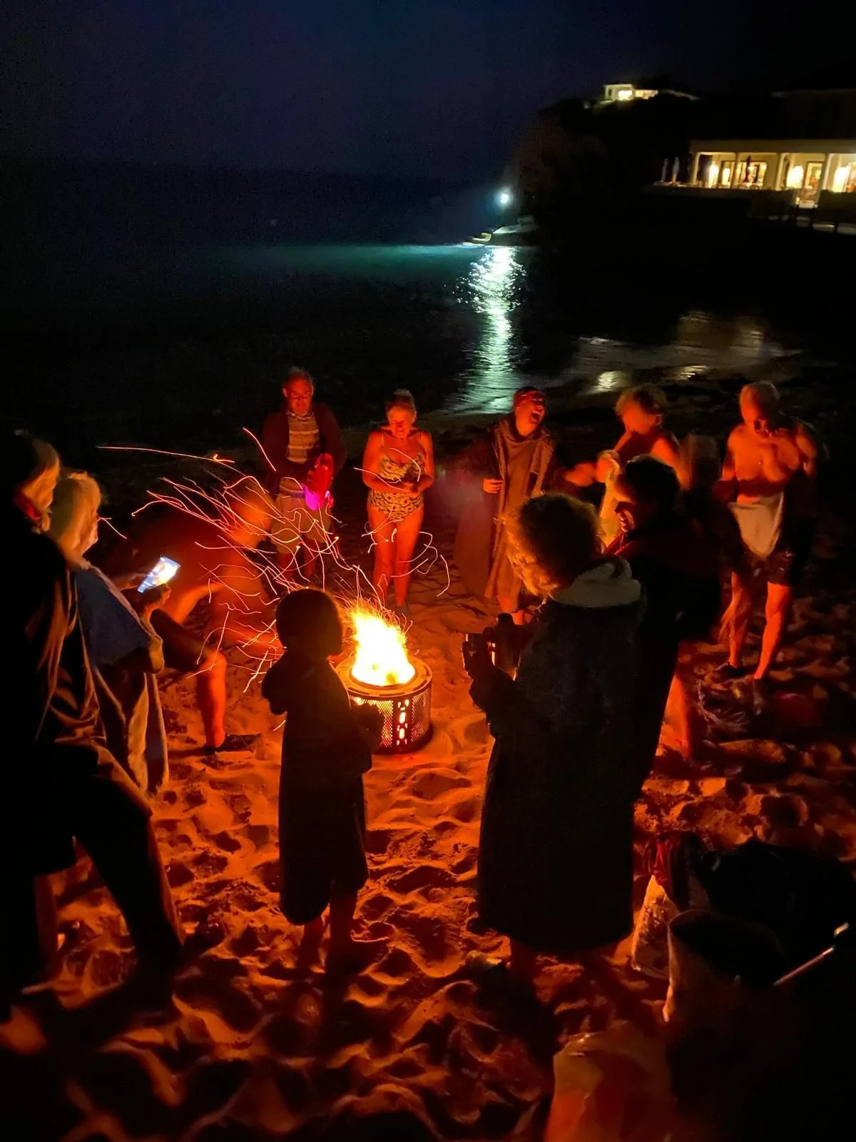 Group of people around a bonfire on the beach at night, some taking photos, with buildings on the shore and a lighthouse in the distance.