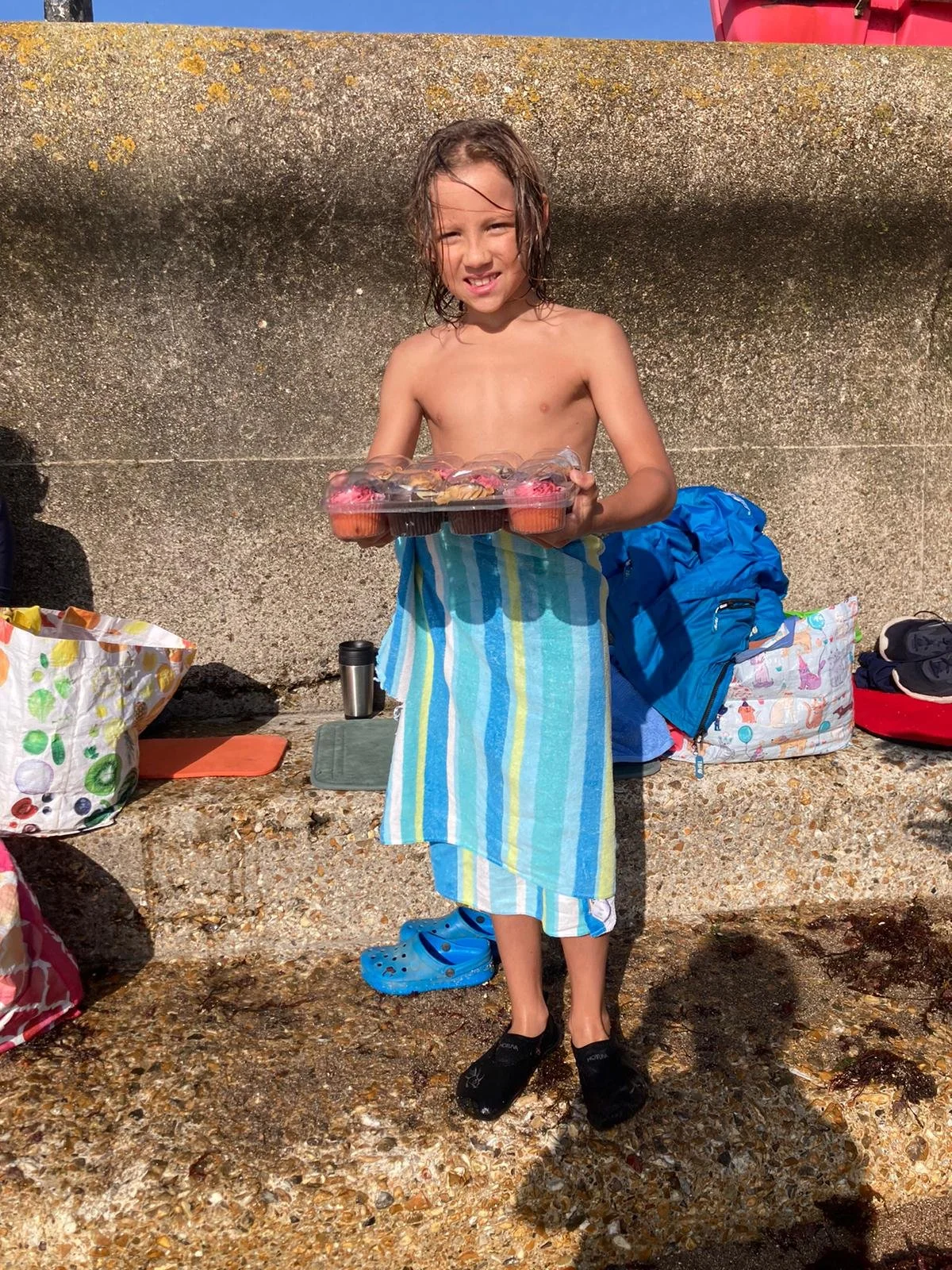 A young girl standing on a gravel beach holding a tray of cupcakes with pink frosting. She is wearing a towel wrapped around her waist, black water shoes, and has wet hair. There are bags and personal items on the ground behind her, and a concrete wa
