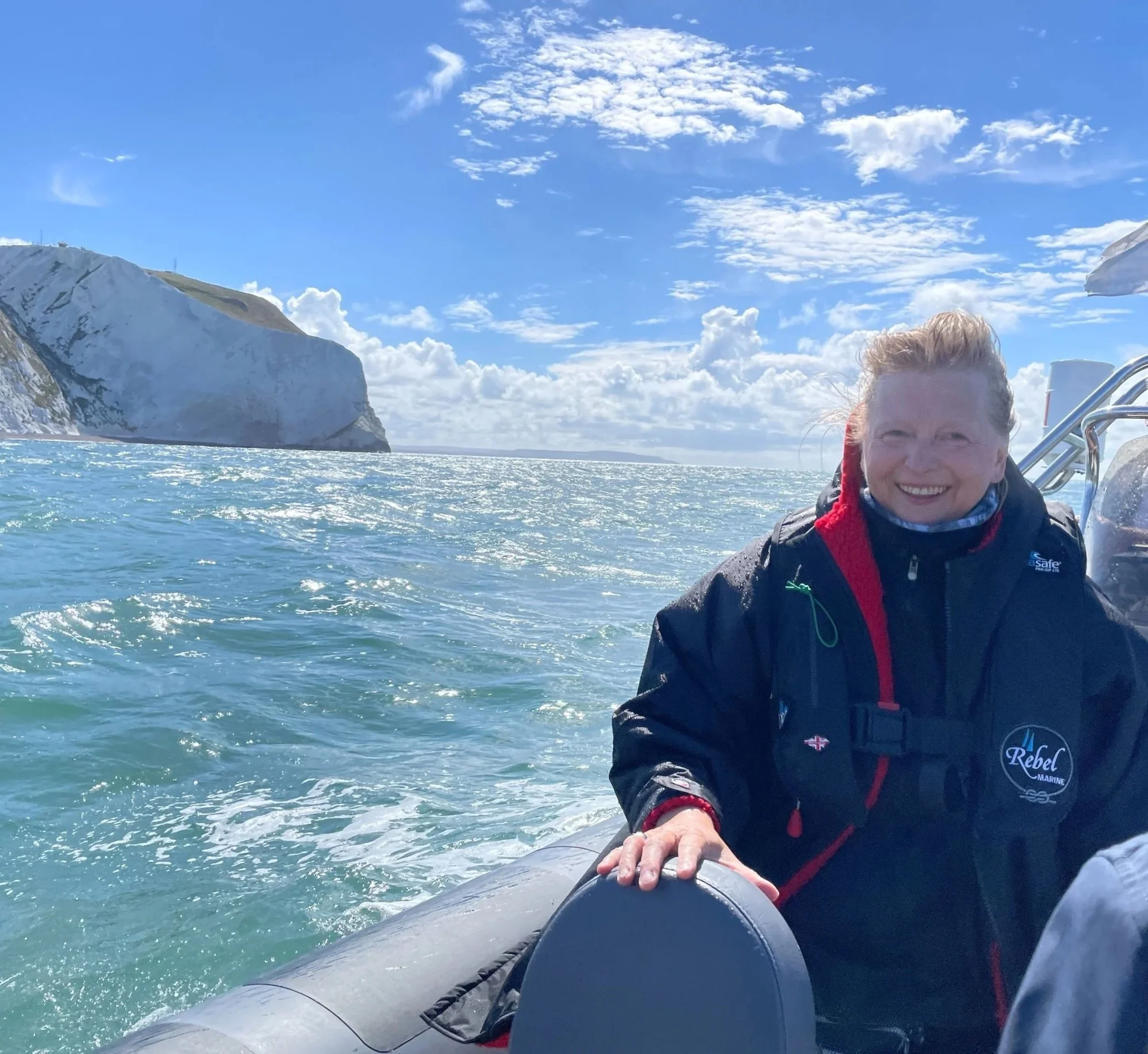 A person with light hair smiling on a boat, with a snow-capped mountain in the background and a partly cloudy blue sky.