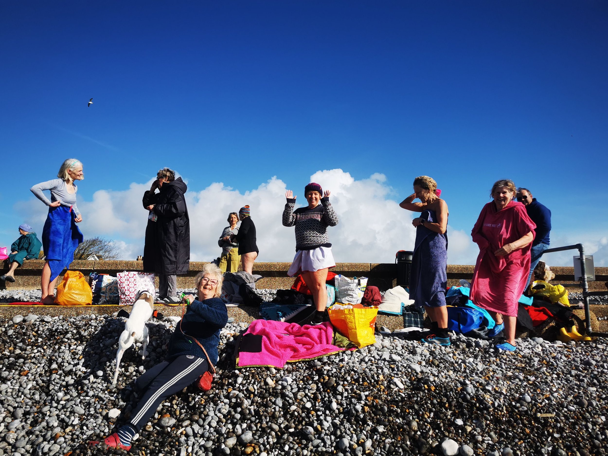 Group of people gathering and chatting outdoors on a rocky beach under a blue sky with clouds, some sitting and some standing, with various bags and blankets around.