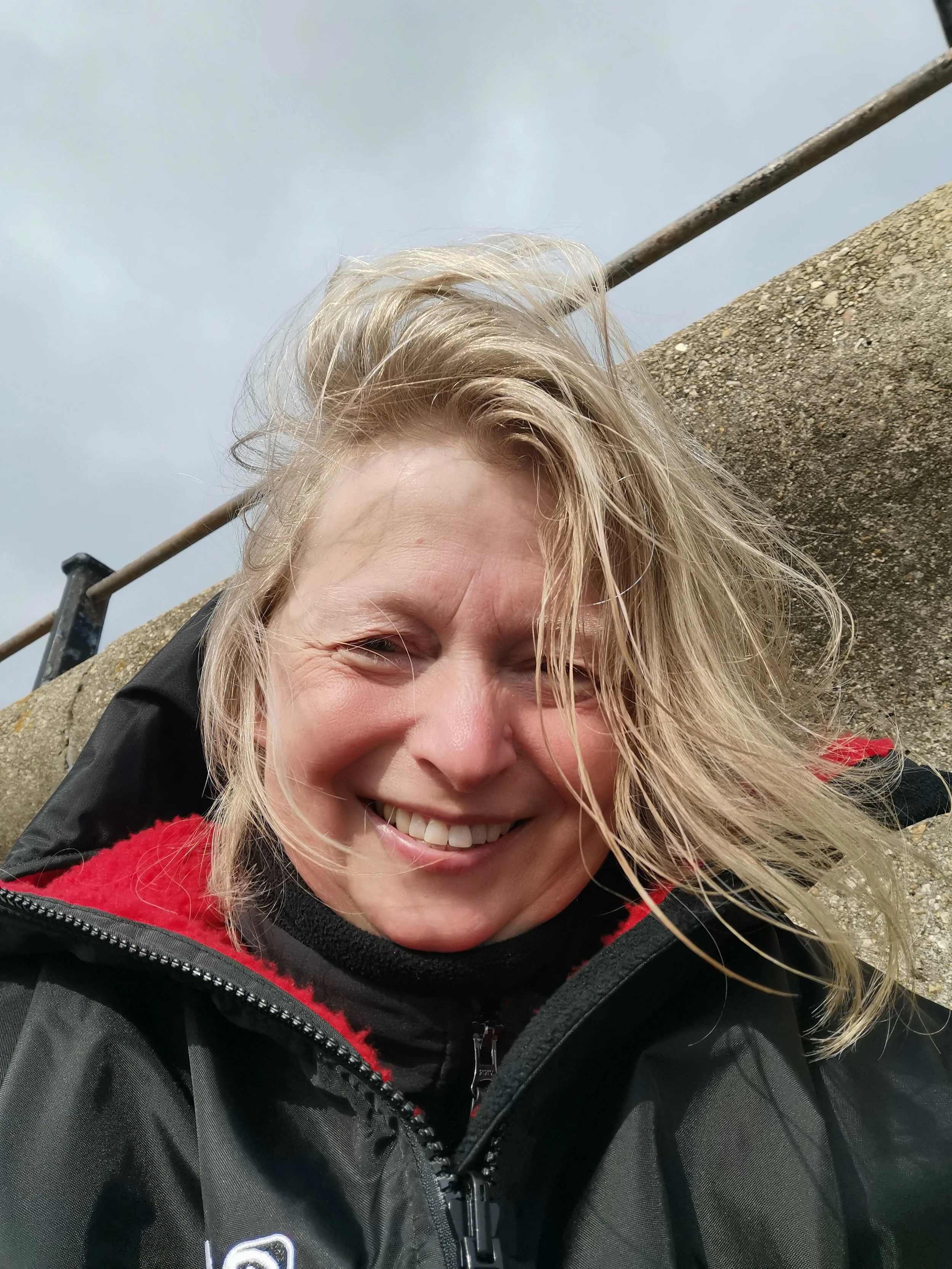 Close-up of a smiling blonde woman with wind-blown hair wearing a black and red outdoor jacket, leaning against a stone surface with a metal railing behind her on a cloudy day.