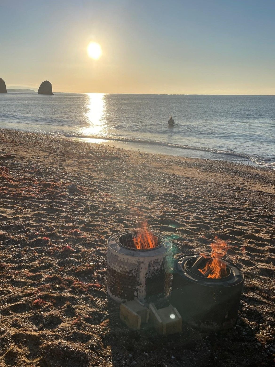 Beach at sunset with two small fires in portable fire pits on the sand, a person wading in the ocean, and large rock formations in the distance.