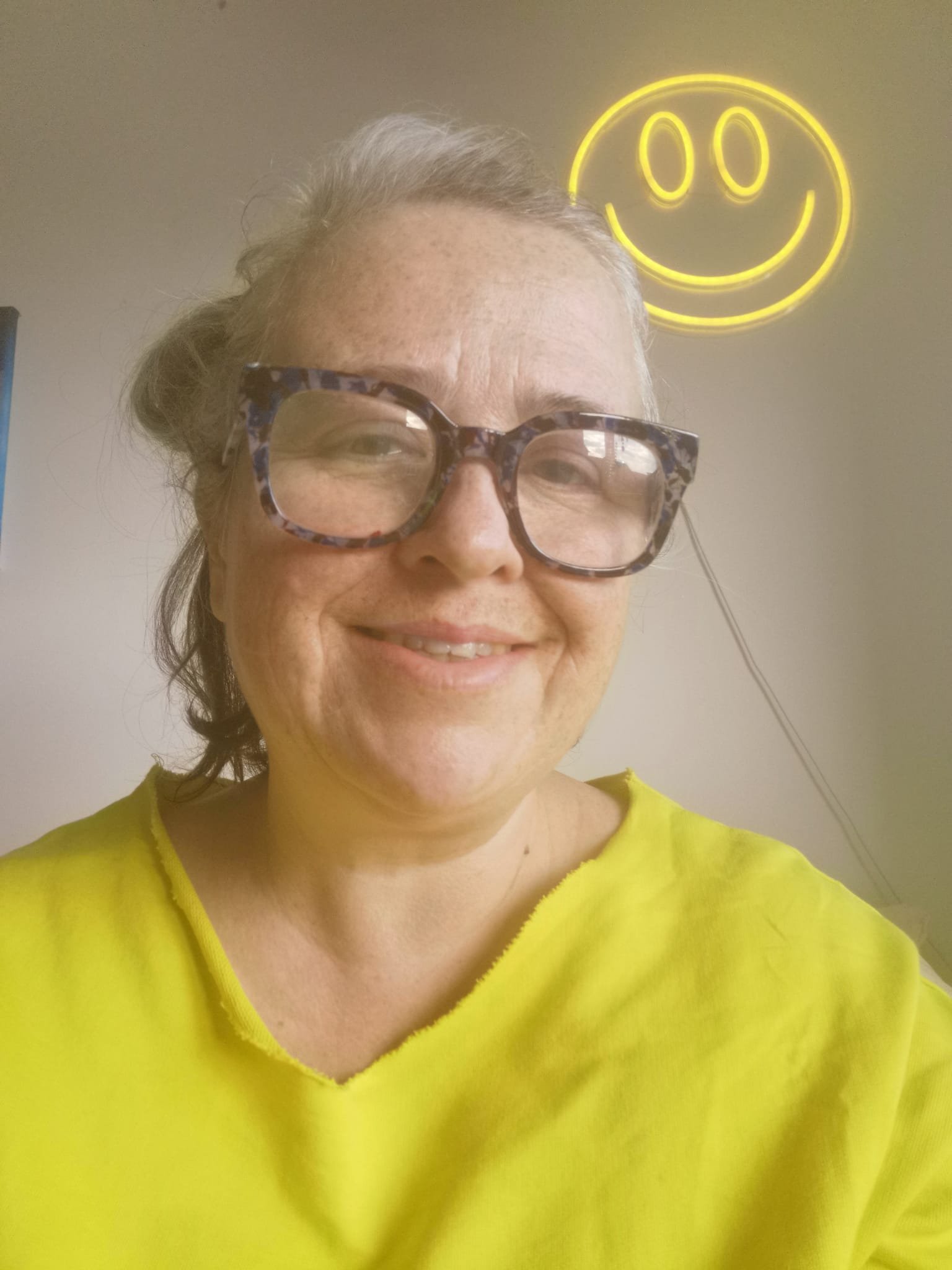 A woman with gray hair wearing leopard print glasses and a yellow top, smiling, with a yellow smiley face neon sign on the wall behind her.