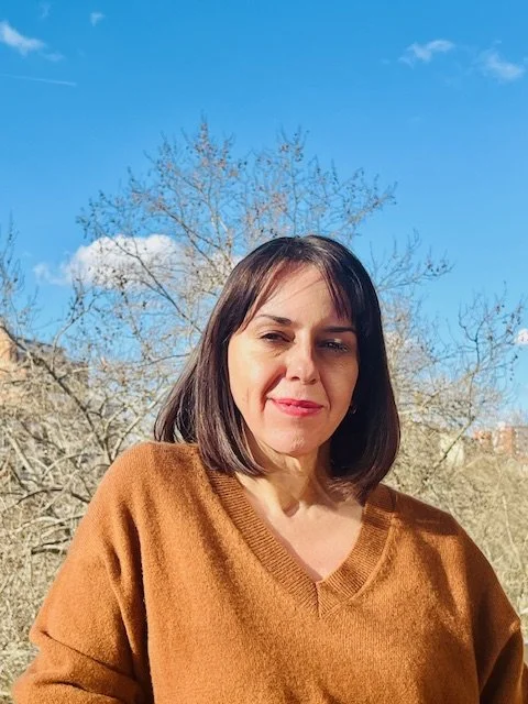 A woman with dark hair and a brown sweater standing outside on a bright day, with a partly cloudy sky and bare trees in the background.