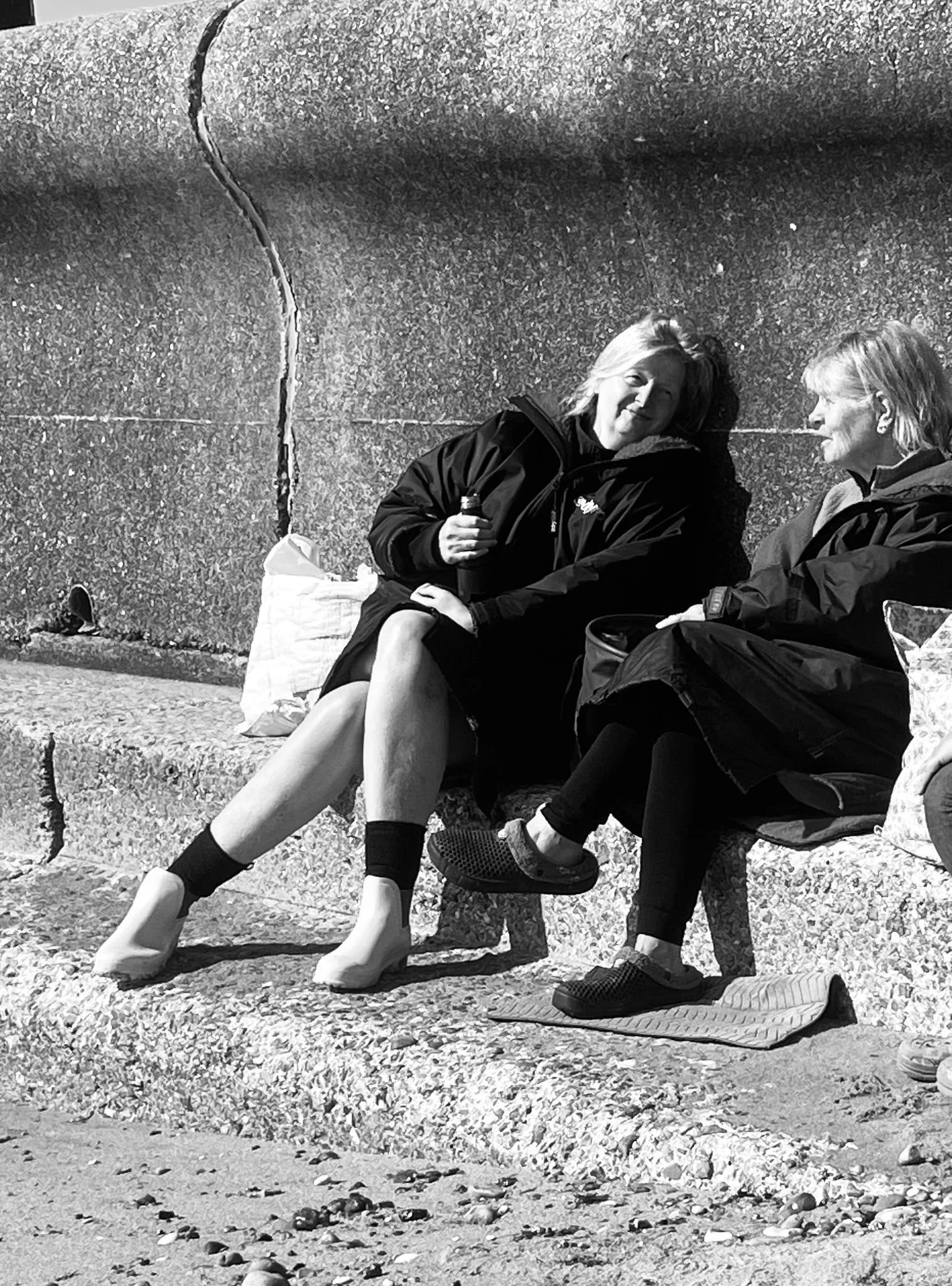 Two women with light hair sitting on a stone barrier at the beach, smiling and talking. One is holding a small bottle, possibly sunscreen or water. They are dressed in casual outdoor clothing, with shoes and bags on the ground near them. The background shows a rocky, textured wall.