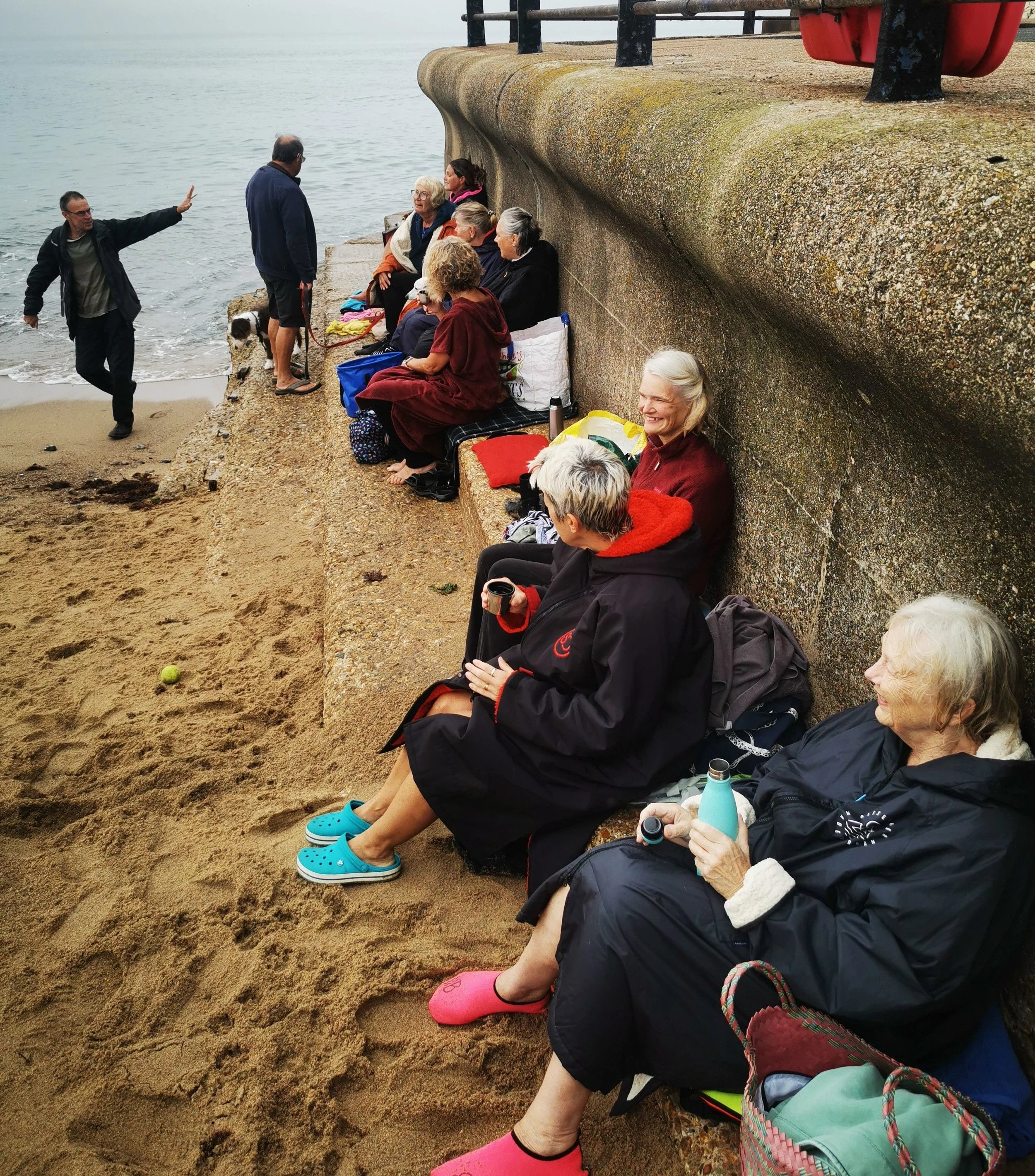 A group of mostly elderly people sitting on a concrete embankment beside a beach, with some wearing colorful rain boots, and a few holding cups or bottles, while a man stands near the water, raising his hand to address them. The scene is overcast, an