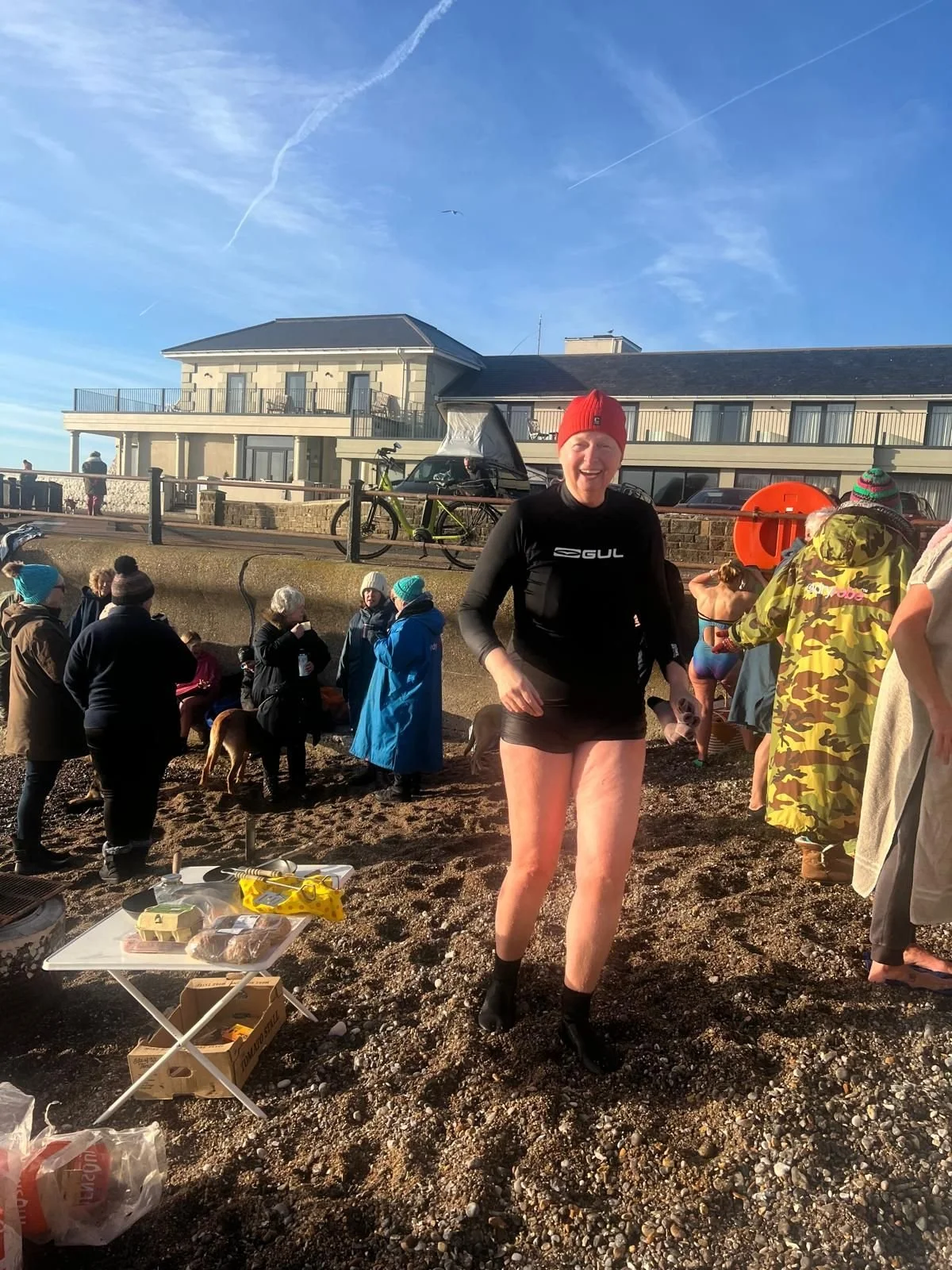A woman with a red hat and black wetsuit smiling on a beach amidst a crowd of people dressed in warm clothing, with a building and bicycles in the background on a clear sunny day.
