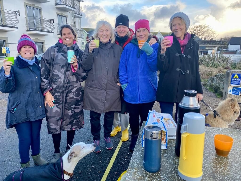 Six women standing outdoors, smiling, holding colorful cups, dressed in winter clothing, with thermoses and a picnic table with snacks and drinks in front of them. Two dogs are present, one on a leash and another lying on the ground. In the background are buildings and partly cloudy sky.