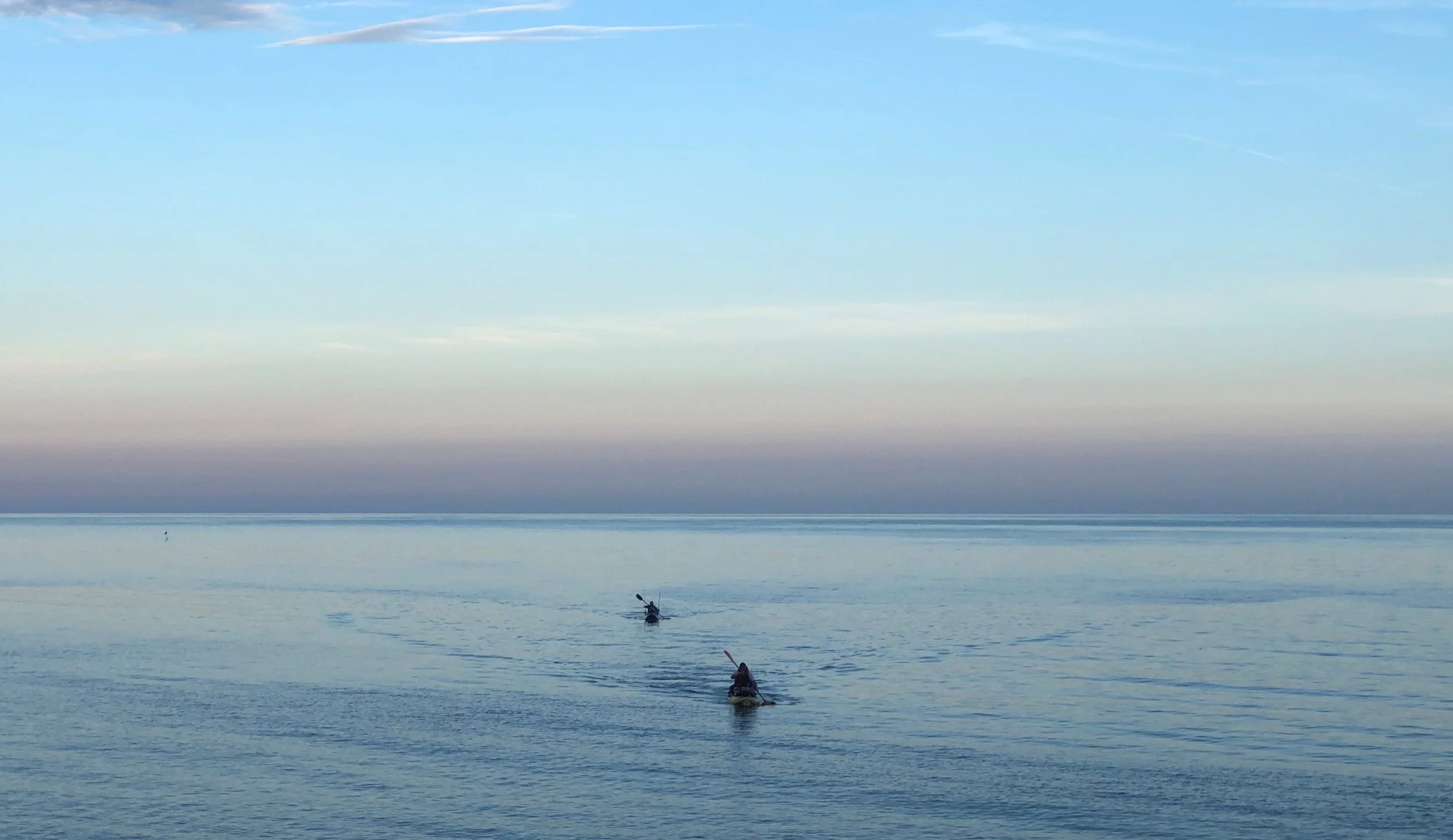 Two people kayaking on calm water with a mostly clear sky and a few wispy clouds above.