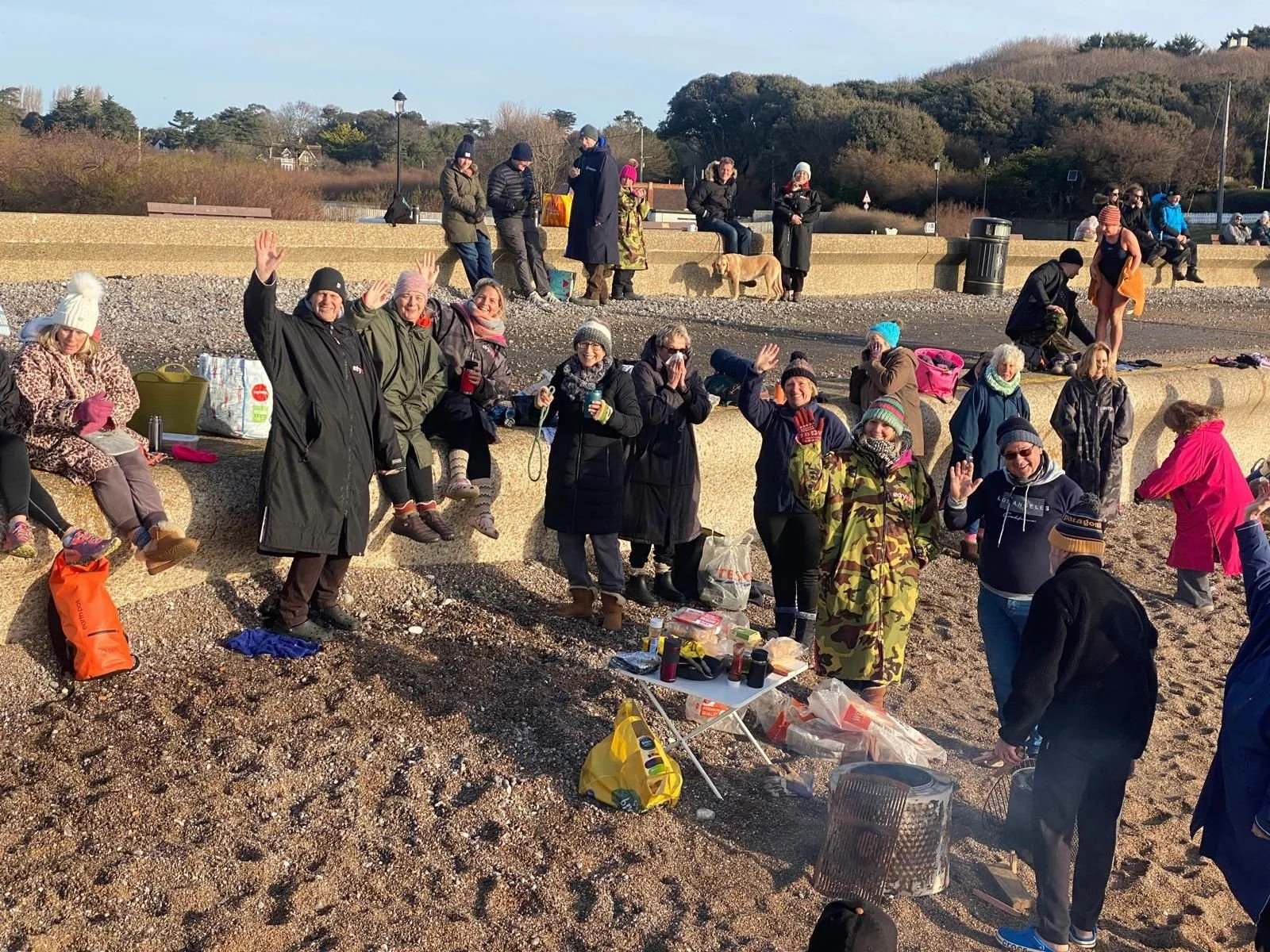 A group of people gathered outdoors on a coastal area with rocks and pebbles, some sitting and others standing, all wearing warm clothing. Some are waving at the camera, and there are tables with food and drinks in the foreground. In the background, 