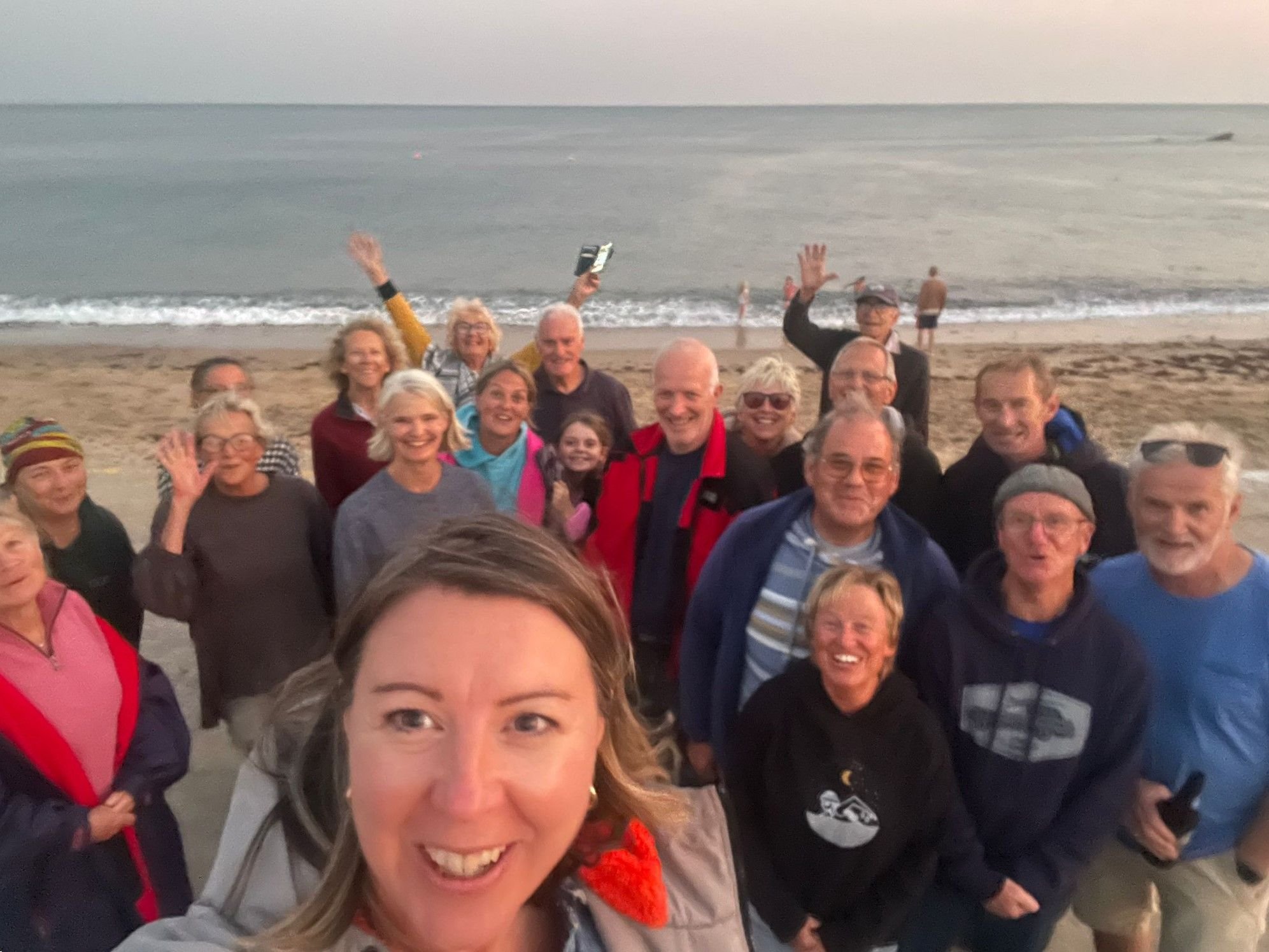 A group of people on a beach taking a selfie, with the ocean in the background. They are smiling, waving, and enjoying their time together.