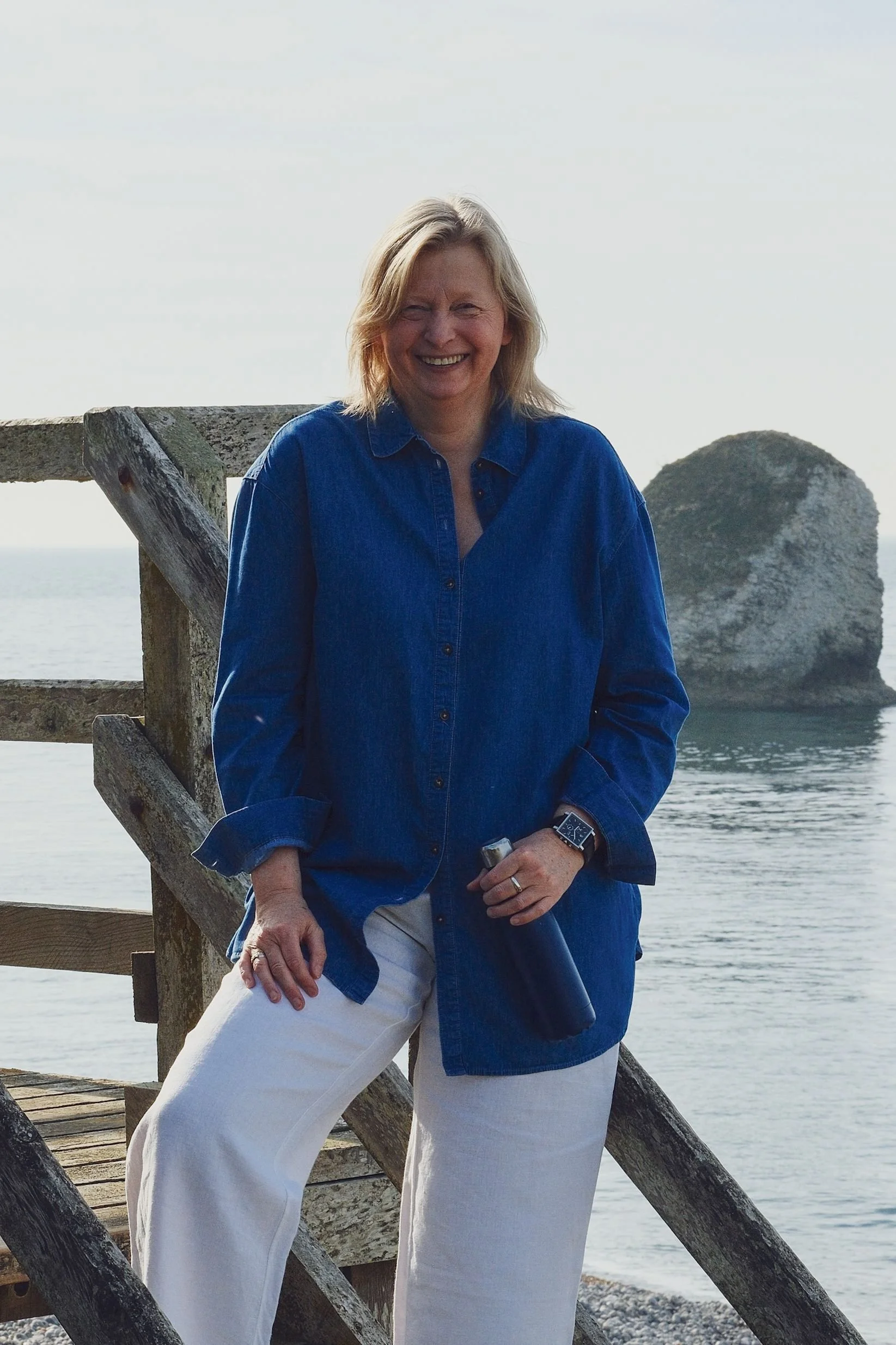 A woman with blonde hair is smiling at the camera, standing outdoors by the water on a wooden dock with a large rock formation in the background. She is wearing a blue denim shirt, beige pants, and a watch.