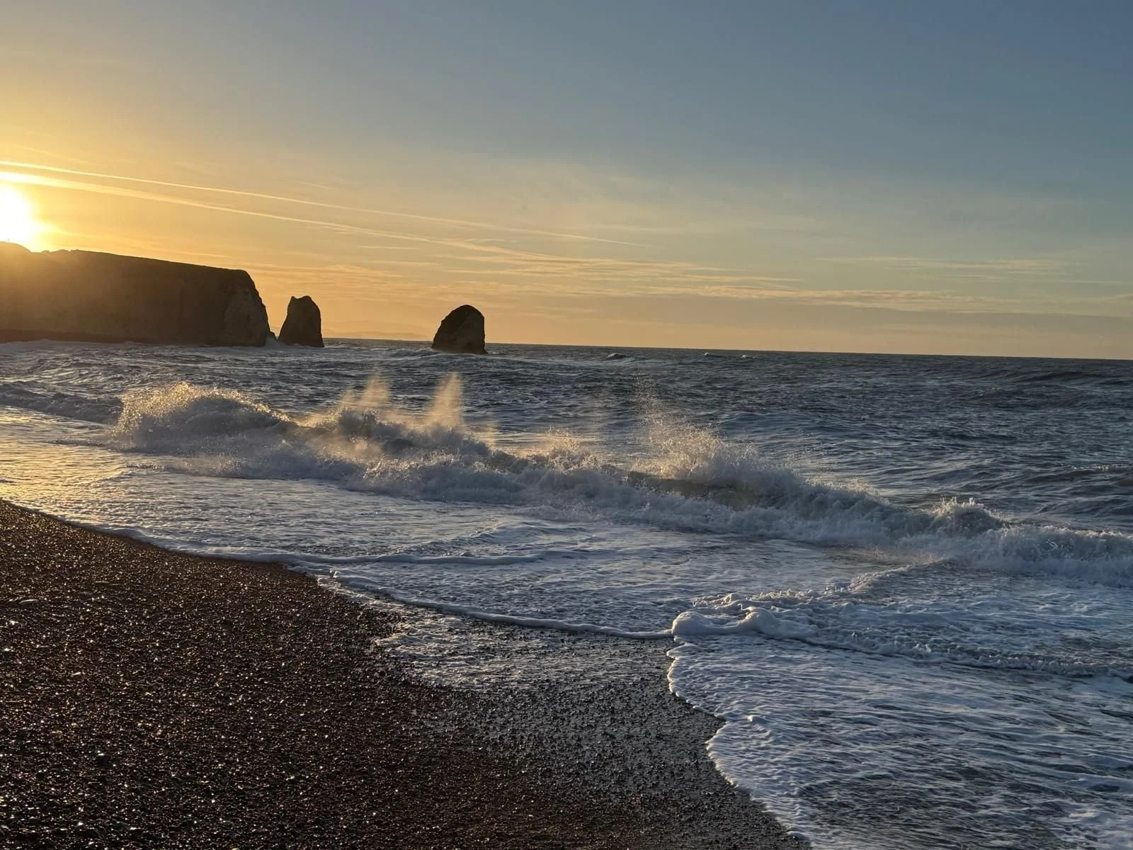 Sunset over a beach with waves crashing and rock formations in the distance.