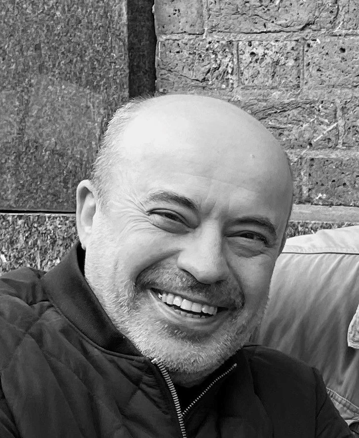 A smiling middle-aged man with a beard, bald head, and light skin, wearing a dark jacket, sitting outside near a brick wall.