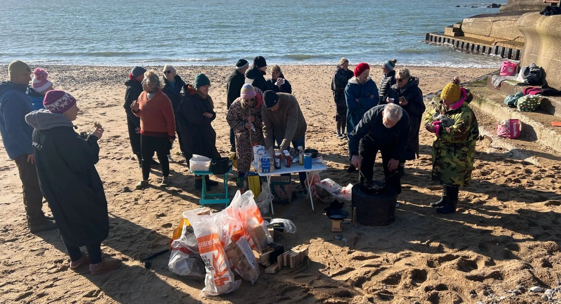 A group of people gathered on a sandy beach near the water, participating in a barbecue or outdoor gathering. Some are standing around a table with food and supplies, others are socializing or cooking.