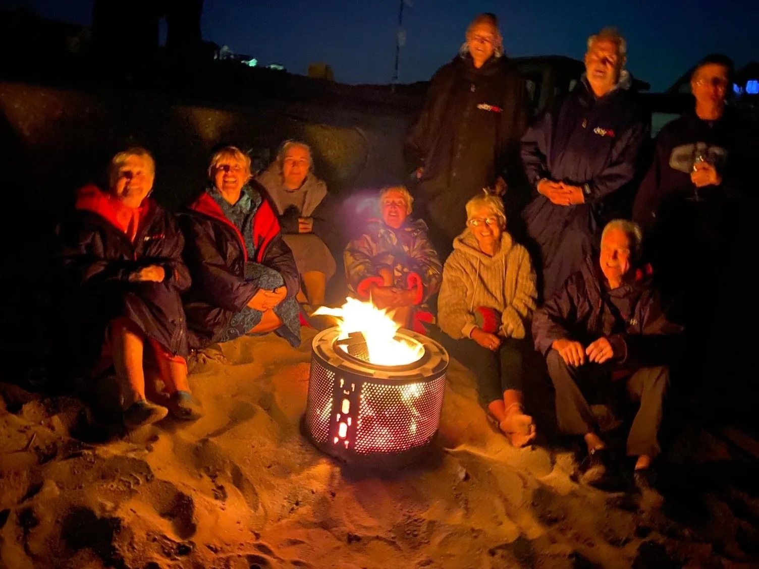 A group of women gathered around a fire pit on a sandy surface at night, smiling and enjoying each other's company.